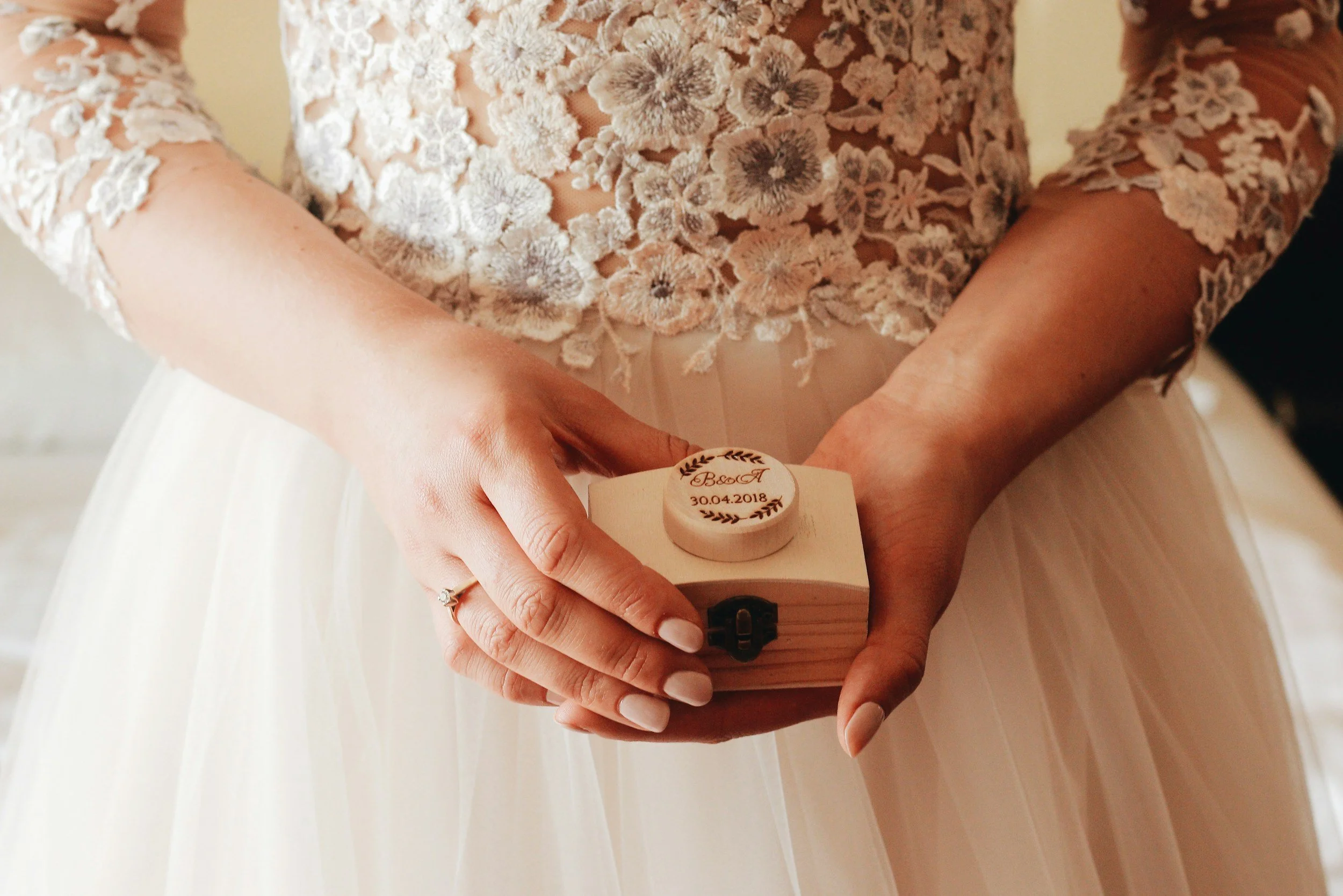 Close-up of a person in a wedding dress holding a small wooden box with a engraved lid. The dress has lace floral details on the sleeves and bodice.