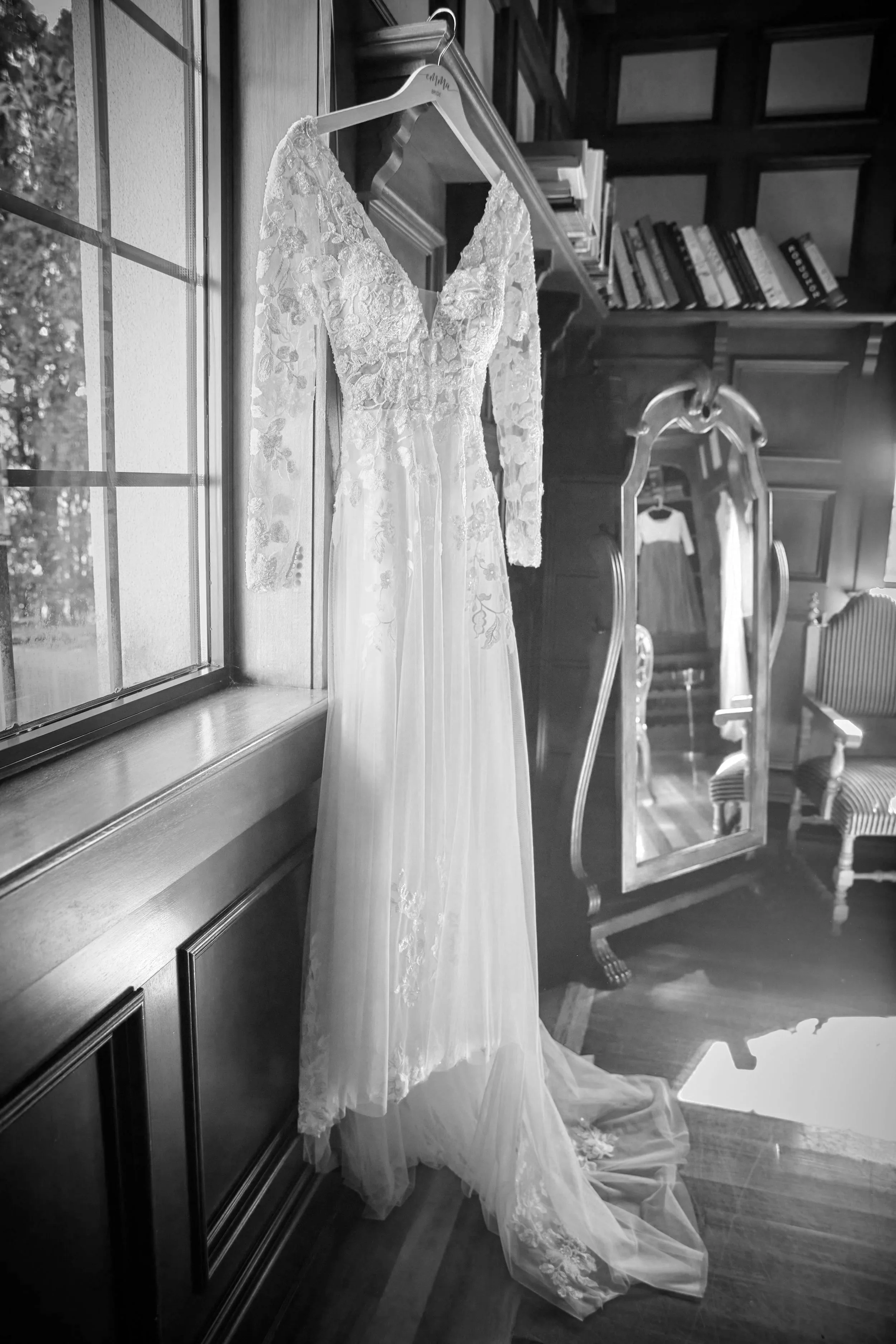 A wedding dress with lace bodice and long lace sleeves hanging on a hanger near a window in a room with wooden furniture and bookshelves.