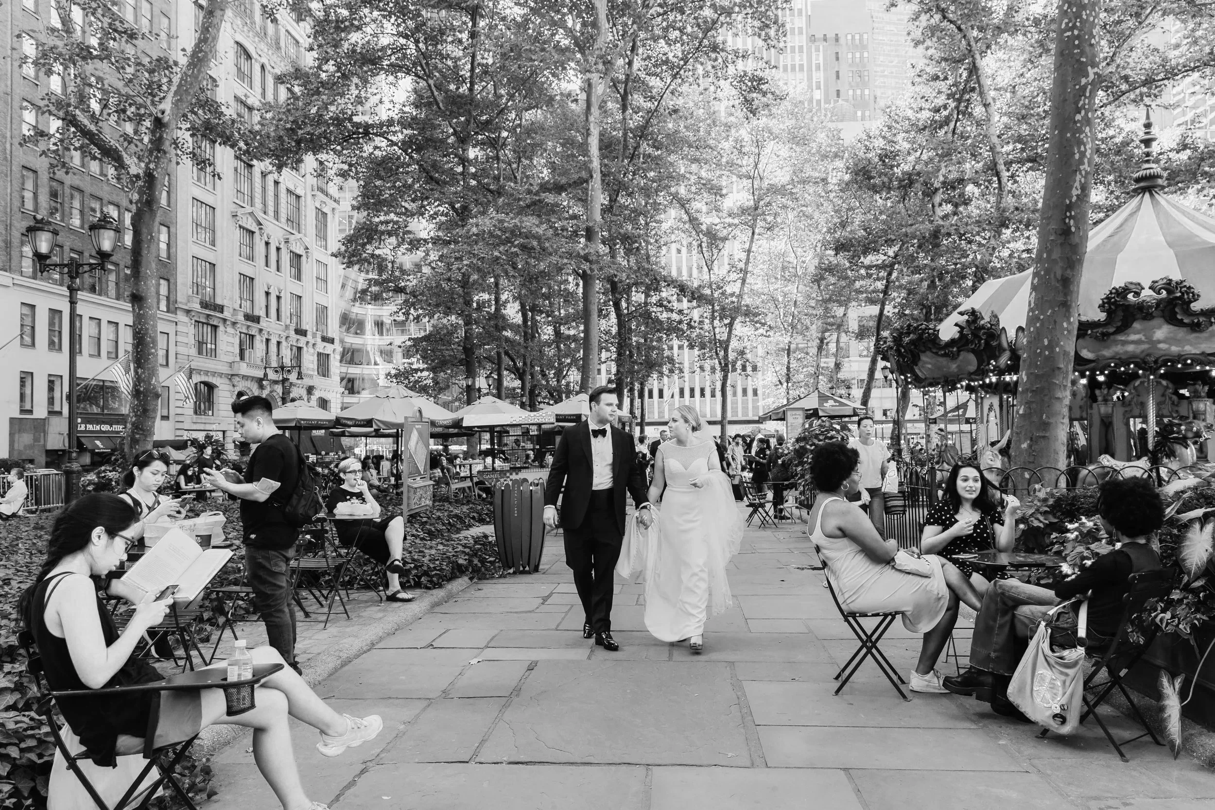 A crowded outdoor scene in a city park with trees, people sitting on benches, and a bride and groom walking hand in hand in wedding attire.