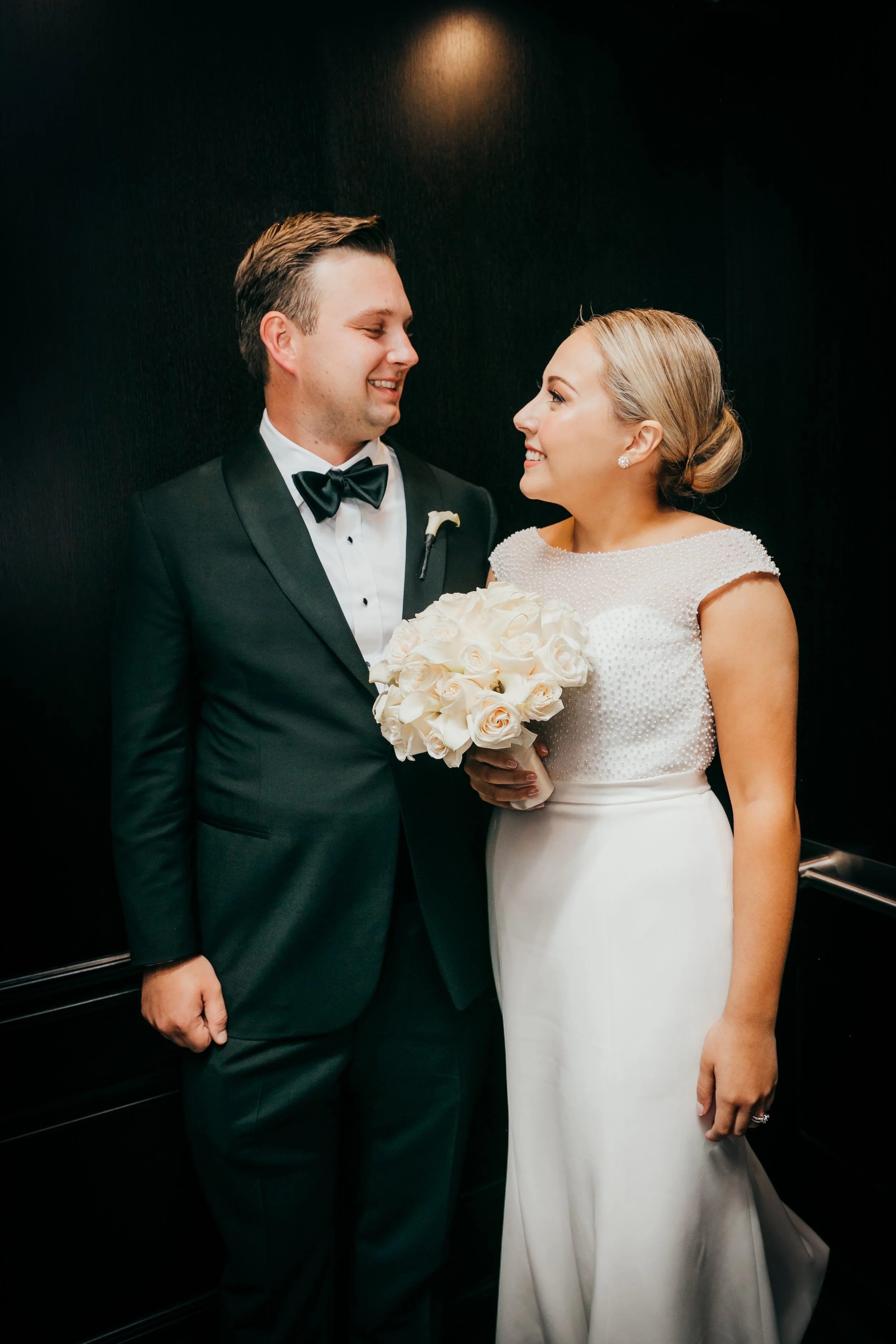 A newlywed couple standing close in an elevator, the groom wearing a black tuxedo with a bow tie and a white shirt, holding a bouquet of white roses, and the bride in a white wedding dress with a beaded bodice, her hair styled in an elegant low bun, and earrings visible.