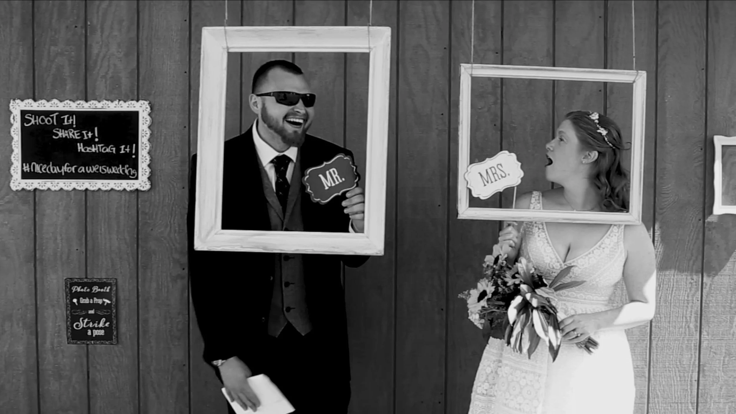 A black and white photo of a newlywed couple standing behind wooden photo frames, with the groom holding a sign that says 'Mr.' and the bride holding a sign that says 'Mrs.' The bride is wearing a wedding dress and holding a bouquet, while the groom is dressed in a suit and wearing sunglasses. They are smiling and appear to be having fun during a wedding photo booth session.