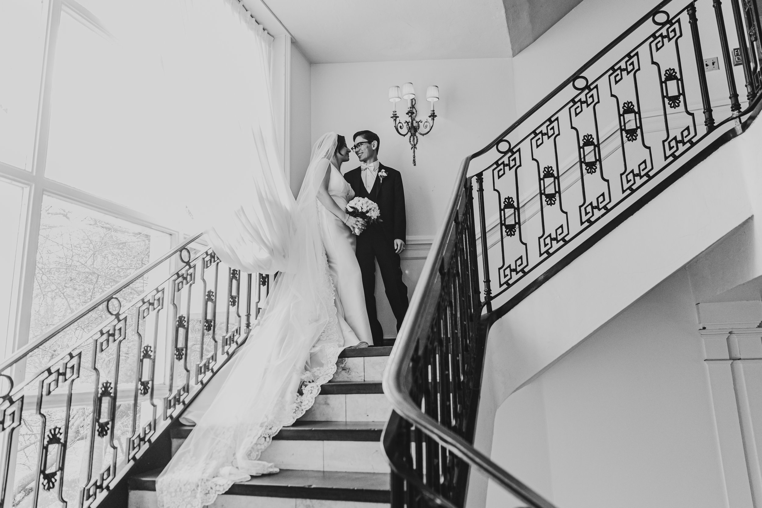 Black and white photo of a bride and groom standing on a staircase indoors. The bride wears a wedding gown with a long train and veil, holding a bouquet of flowers. The groom wears a black tuxedo with a bow tie and glasses. They are smiling and looking at each other. A decorative wall sconce with three lamps is on the wall behind them, and a window with curtains is visible to the left.