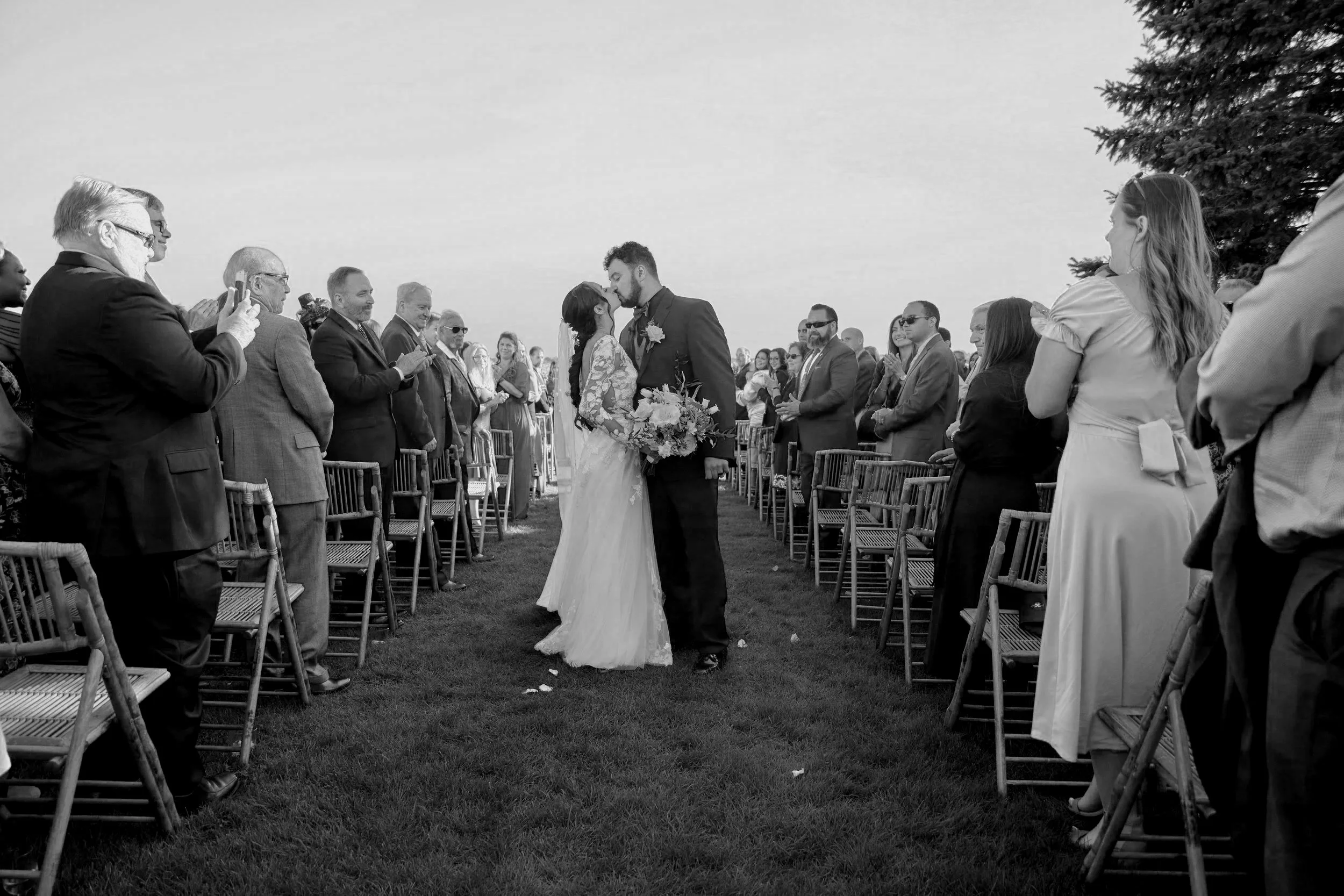 A black and white photo of a wedding ceremony outdoors with a bride and groom kissing, surrounded by seated guests.