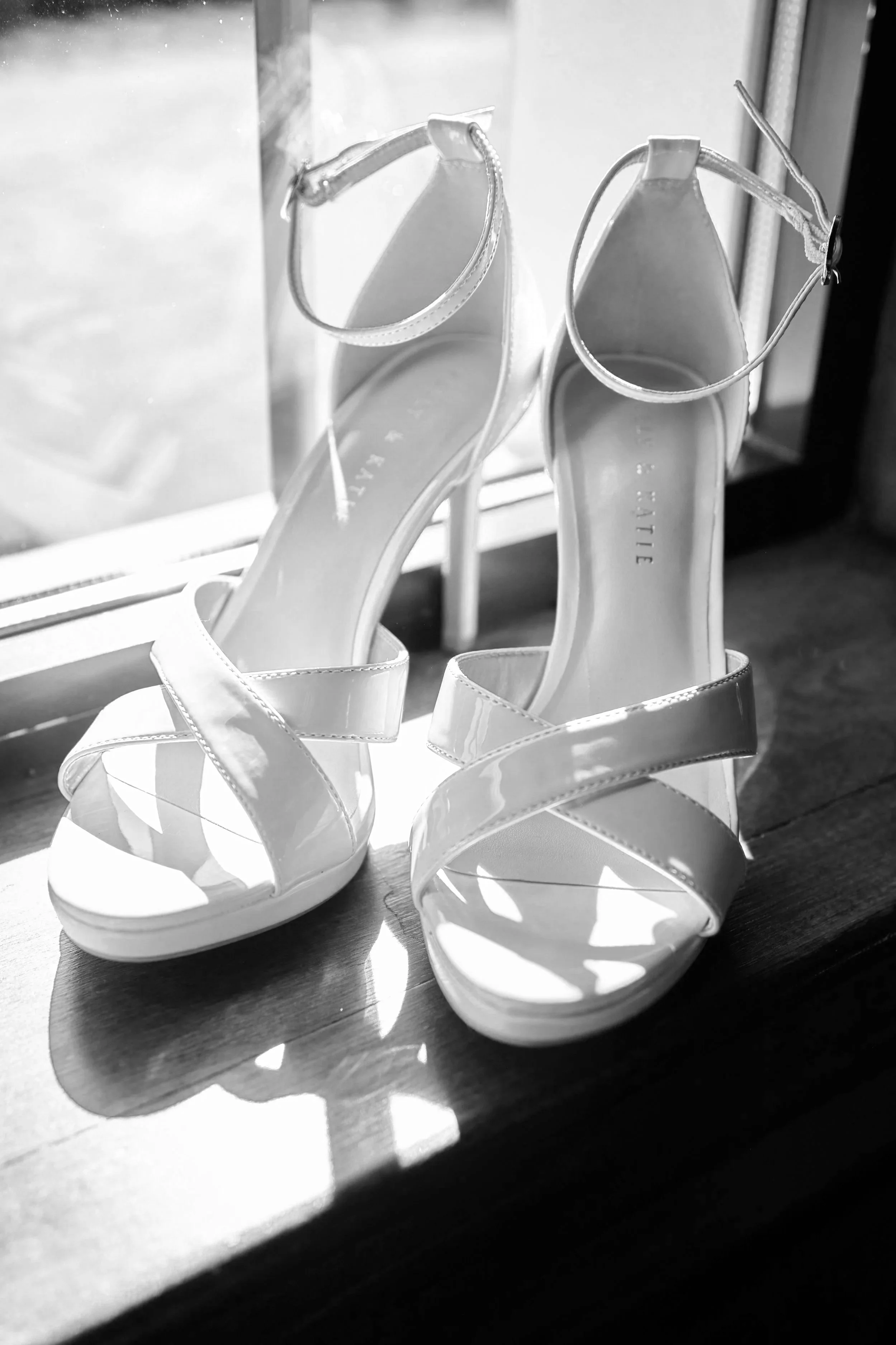 A pair of white high-heeled shoes with ankle straps sitting on a wooden surface near a window, casting shadows.