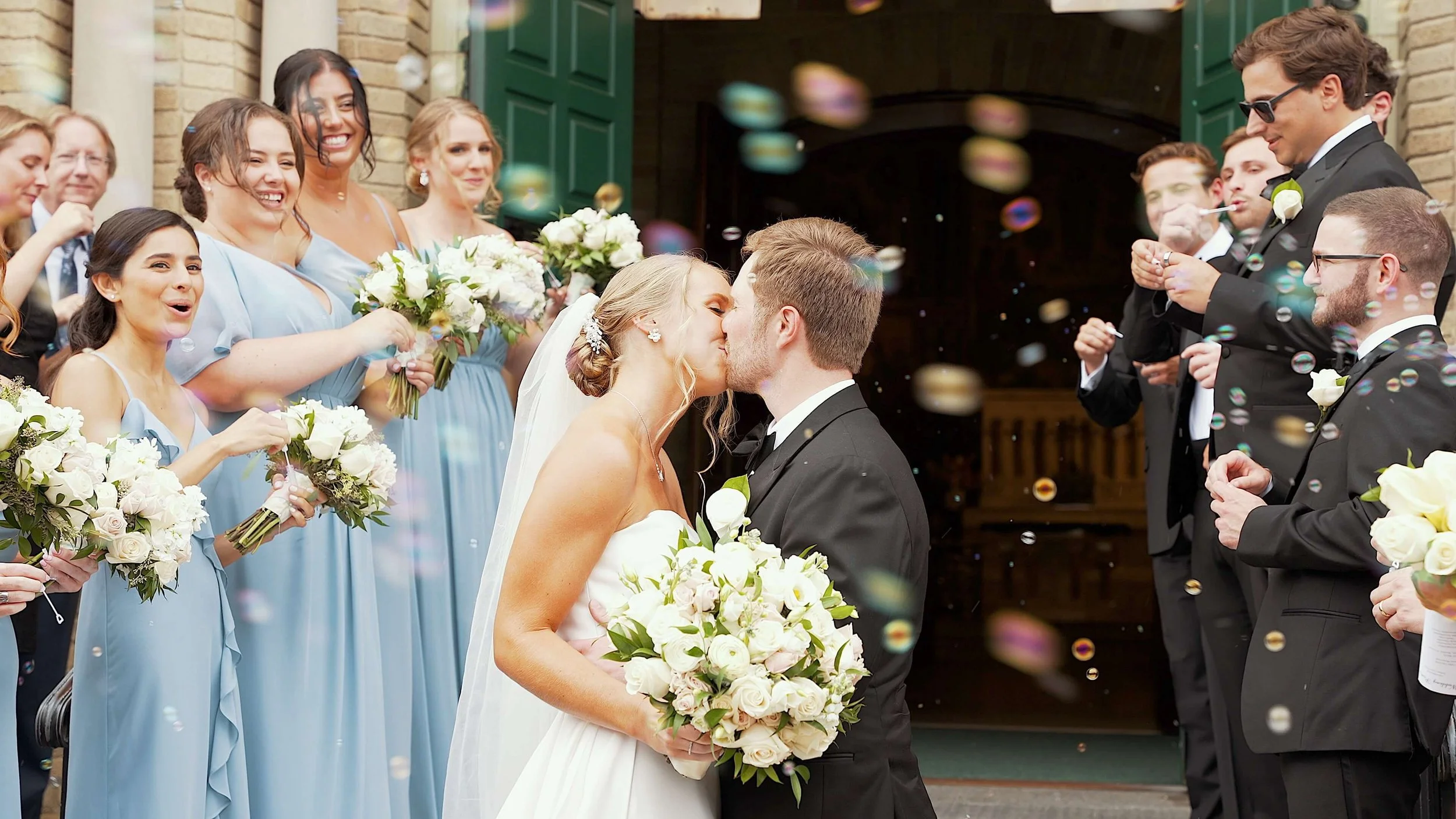 A bride and groom kiss as they start their wedding, surrounded by bridesmaids in light blue dresses and groomsmen in black tuxedos outside a church, with confetti falling around them.