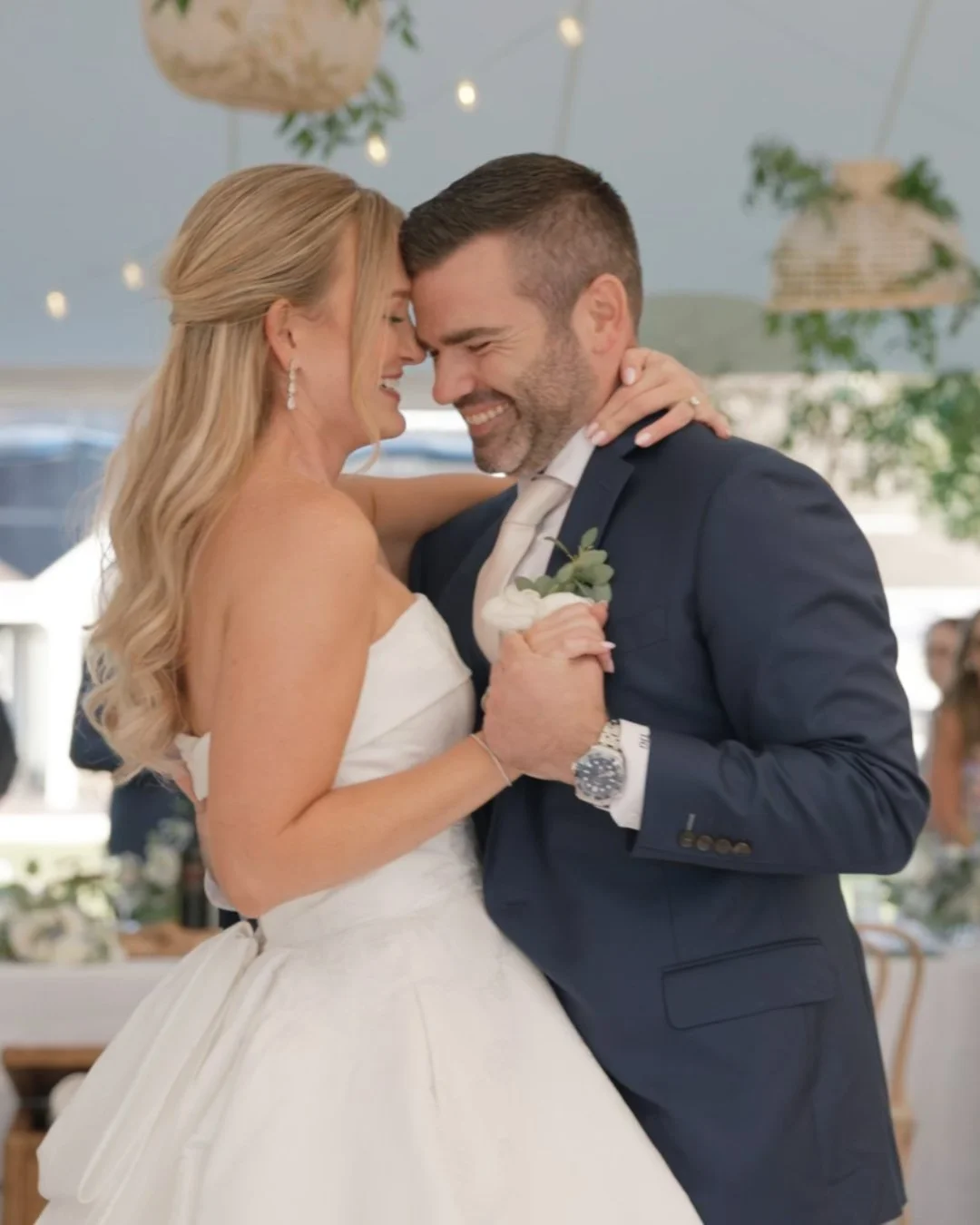 A bride and groom sharing a dance or moment at their wedding, touching foreheads and smiling in an indoor setting with hanging lights and plants.