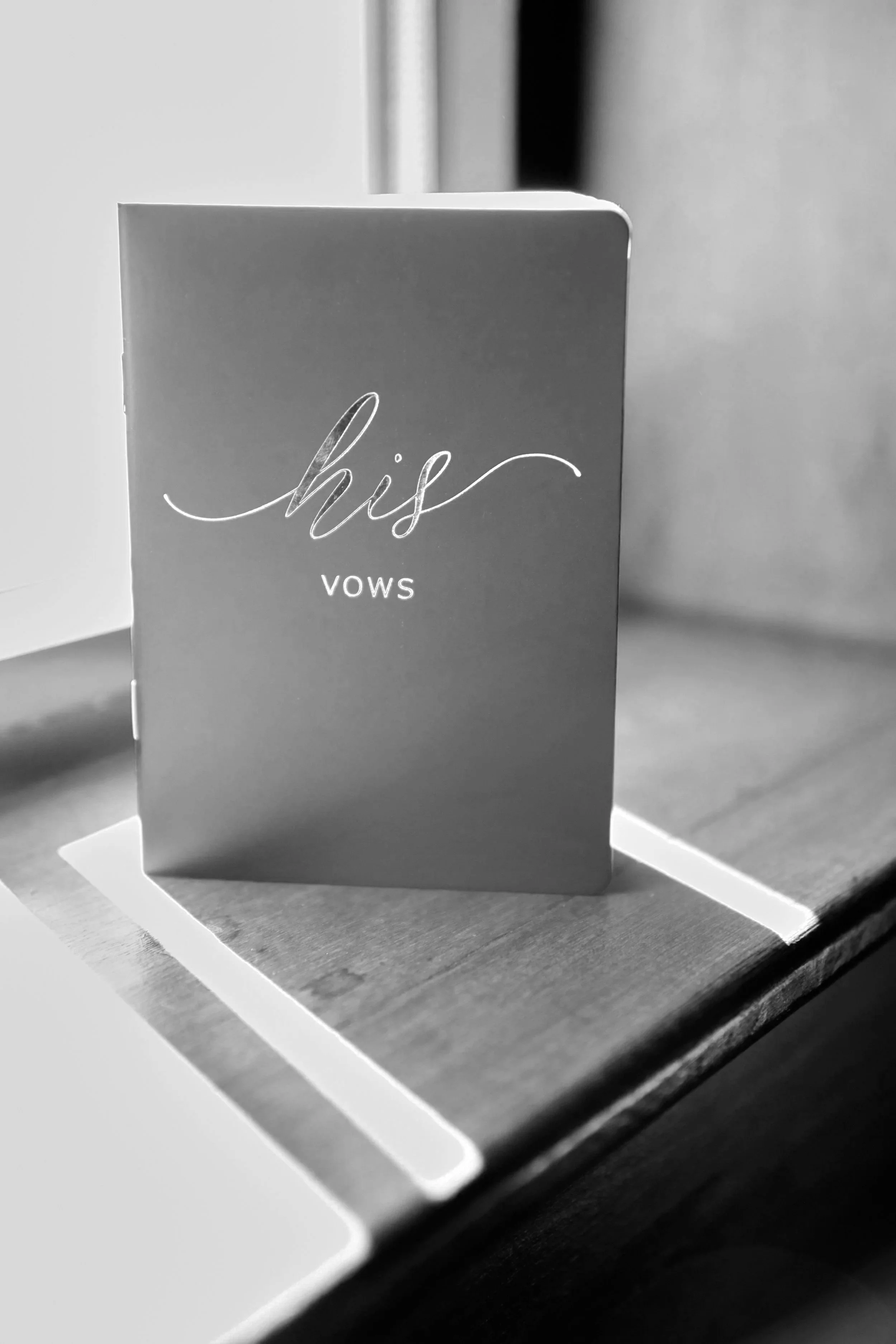 A greeting card standing on a desk with the words 'his vows' written on the front, in front of a white RSVP card.