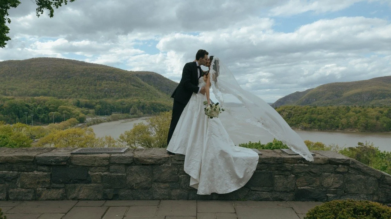 A bride and groom share a kiss on a stone ledge with scenic hills and a river in the background under cloudy skies.