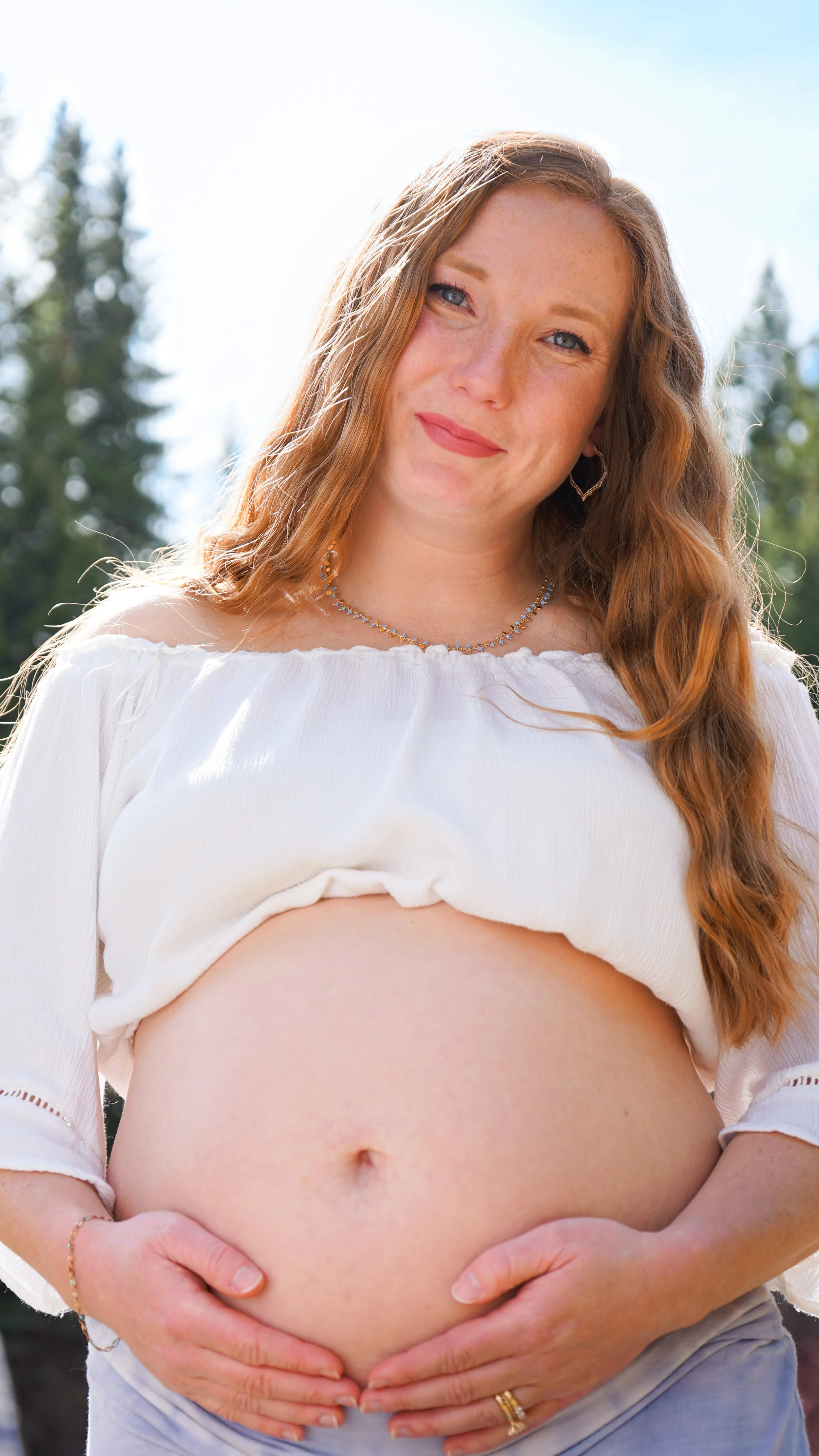 A pregnant woman with long reddish hair, wearing a white off-shoulder top and jewelry, standing outdoors with a background of trees and a bright sky, holding her pregnant belly.