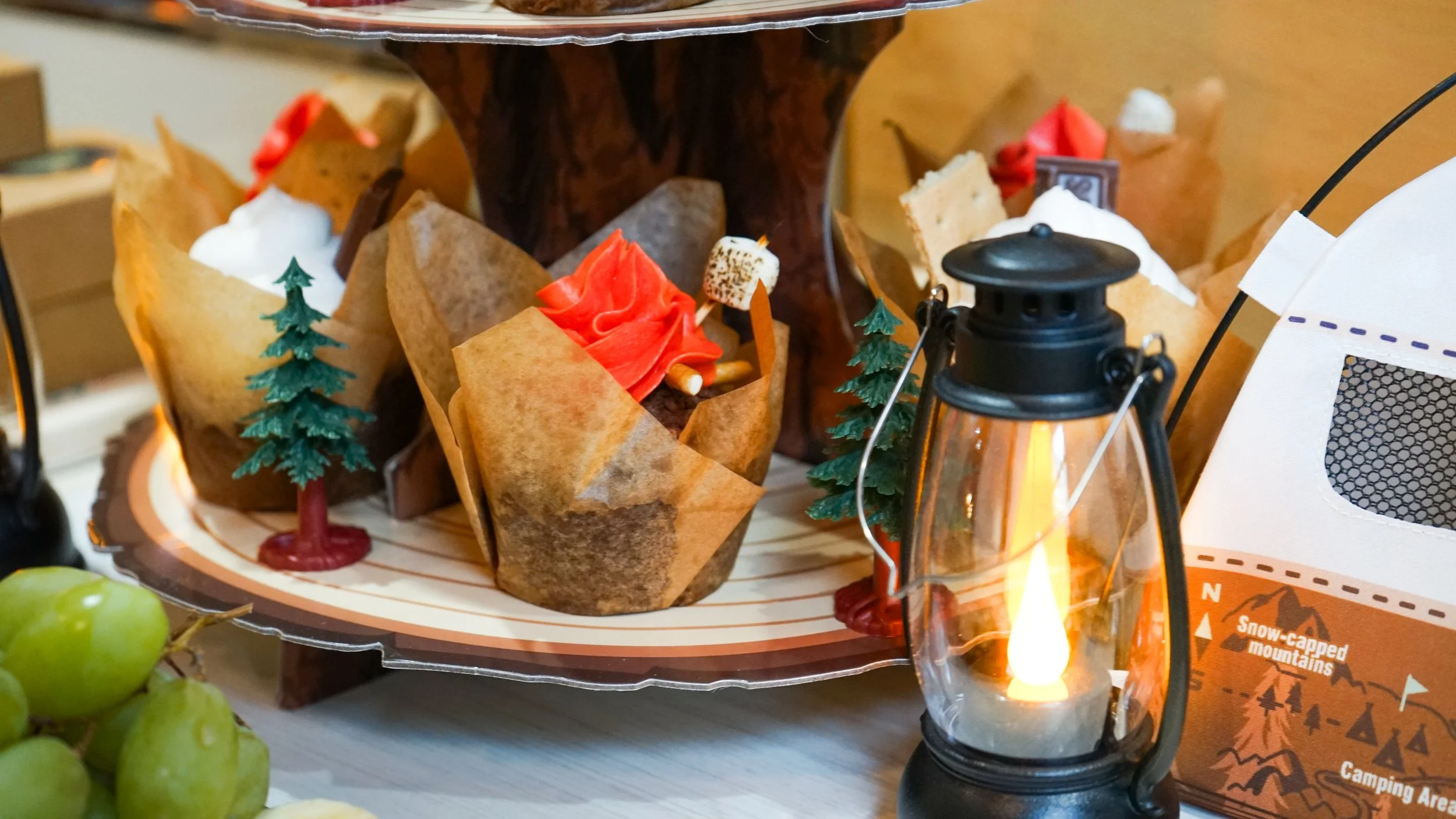 Decorative camping-themed display with miniature trees, a lantern, marshmallows, and a red fabric resembling a campfire, surrounded by paper and plastic elements.