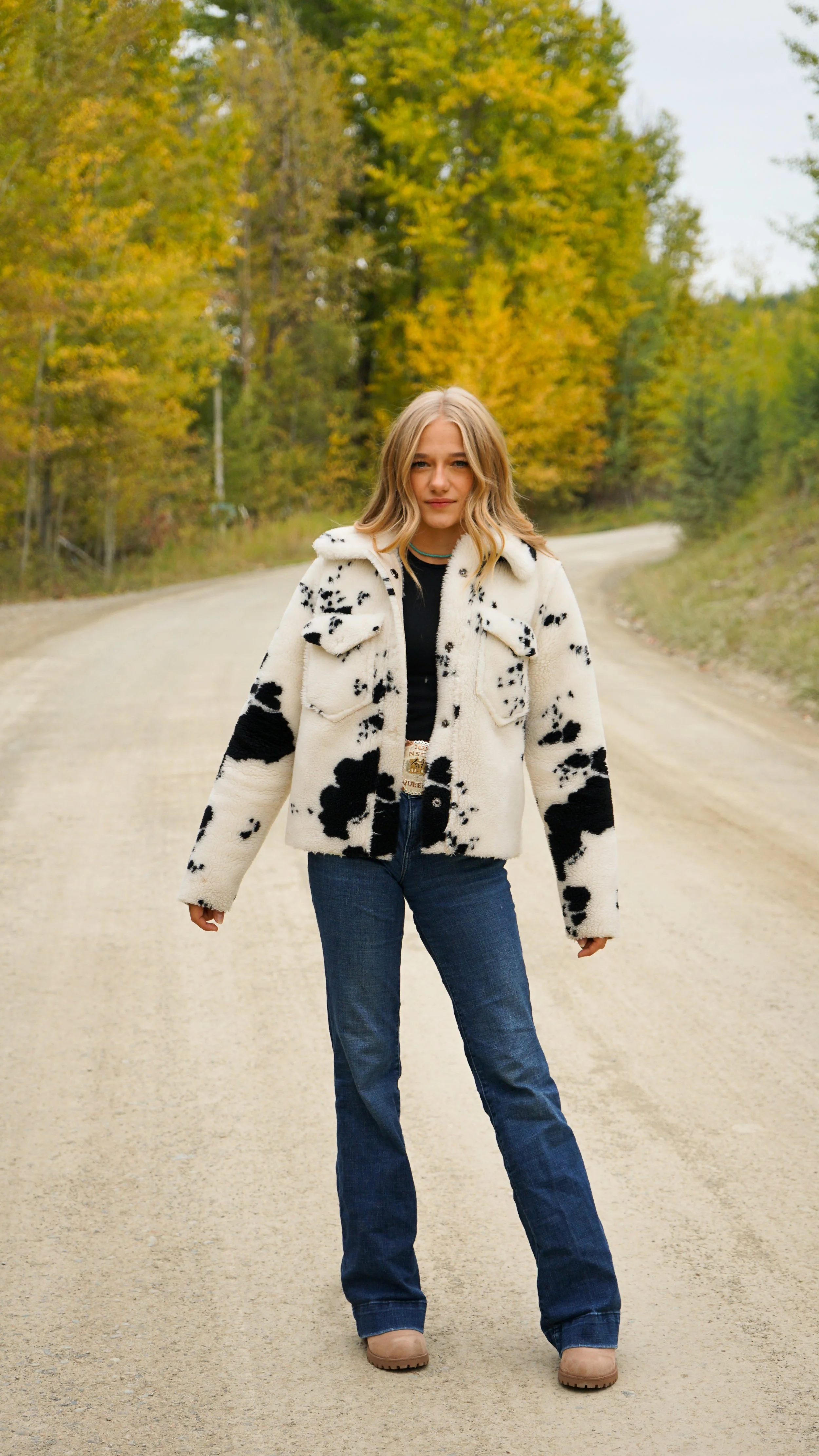 A woman with blonde hair wearing a white and black cow print jacket, black top, blue jeans, and tan boots standing on a dirt road with autumn trees in the background.