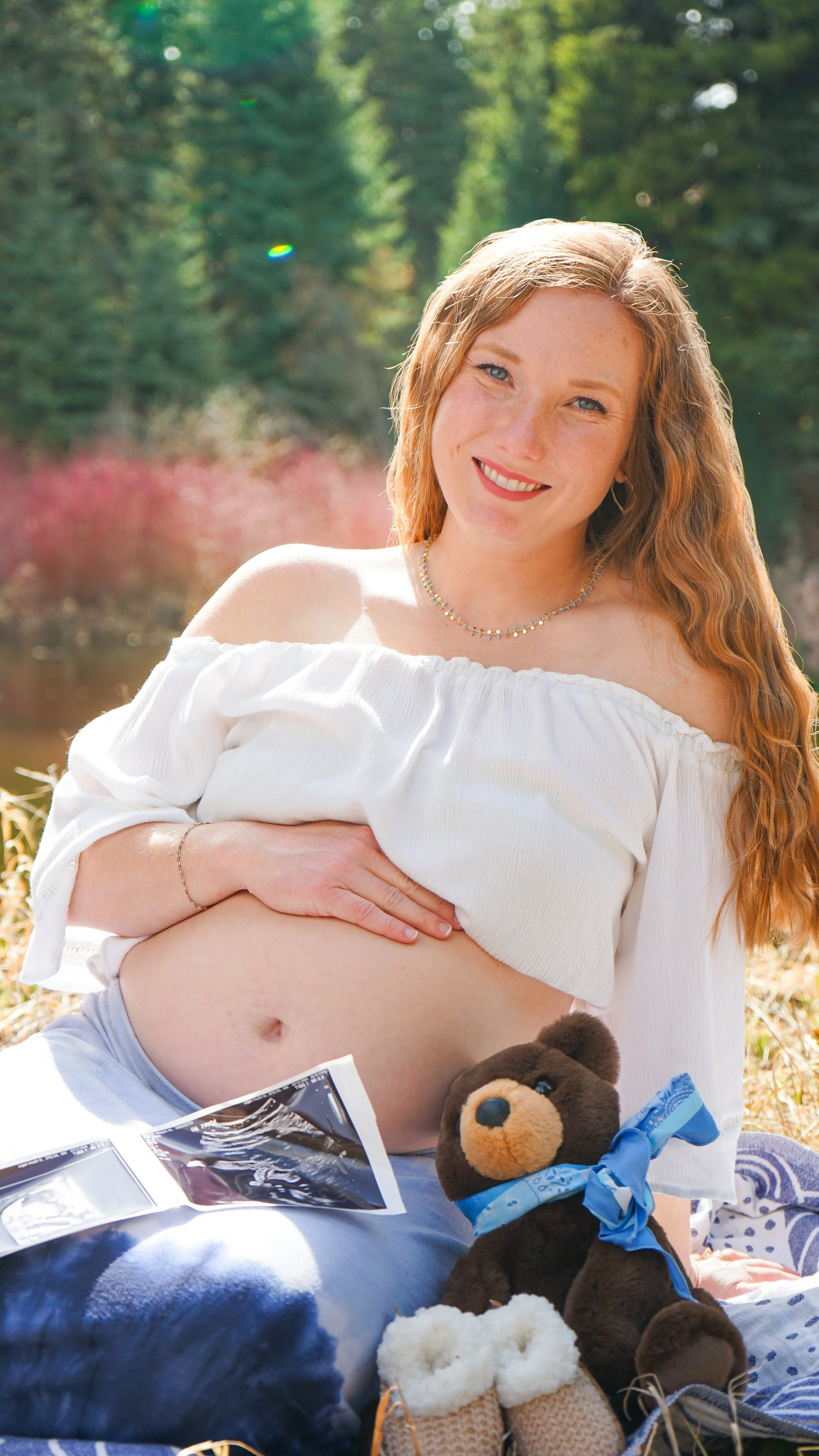 A smiling woman with long red hair, wearing an off-shoulder white top, outdoors in a natural setting with trees and sunlight, holding her belly, with ultrasound images, a teddy bear, a pair of baby shoes, and other baby items in front of her.