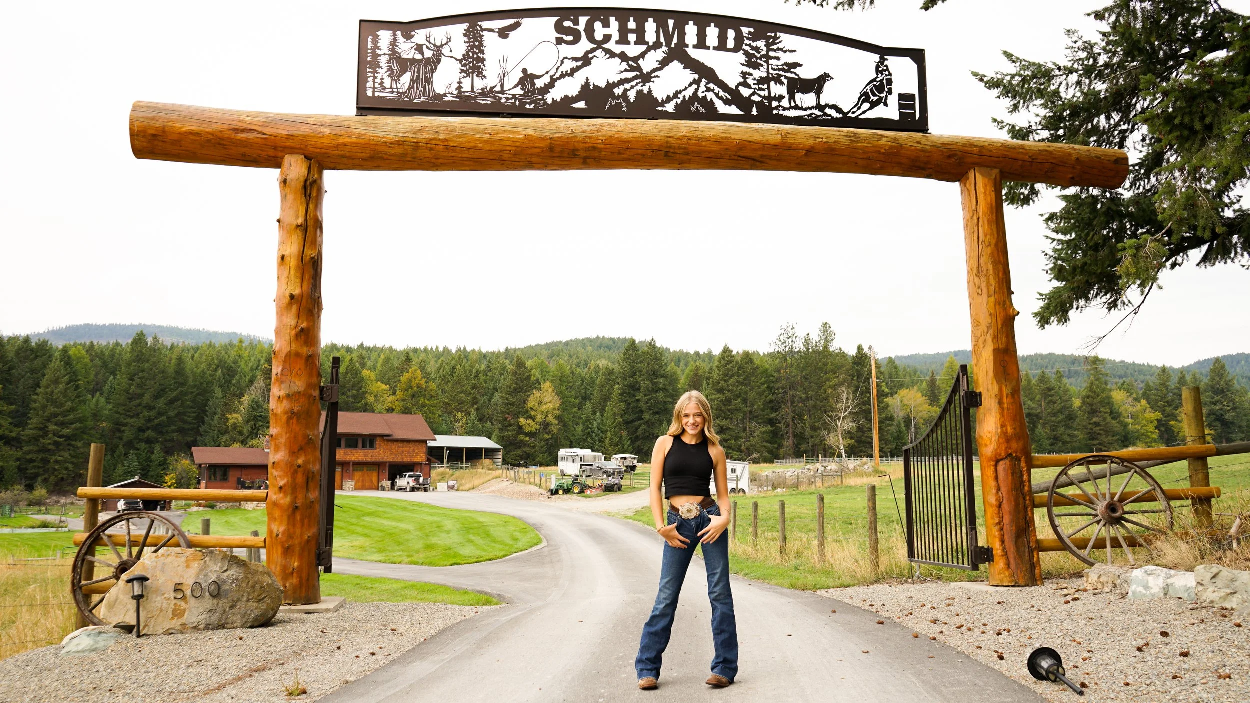 A woman with long blonde hair, wearing a black sleeveless top and jeans, standing in front of a large wooden gate with a mountain scene and the word "Schmidt" on top. The gate is made of logs and is supported by two wooden posts. There are trees, bui