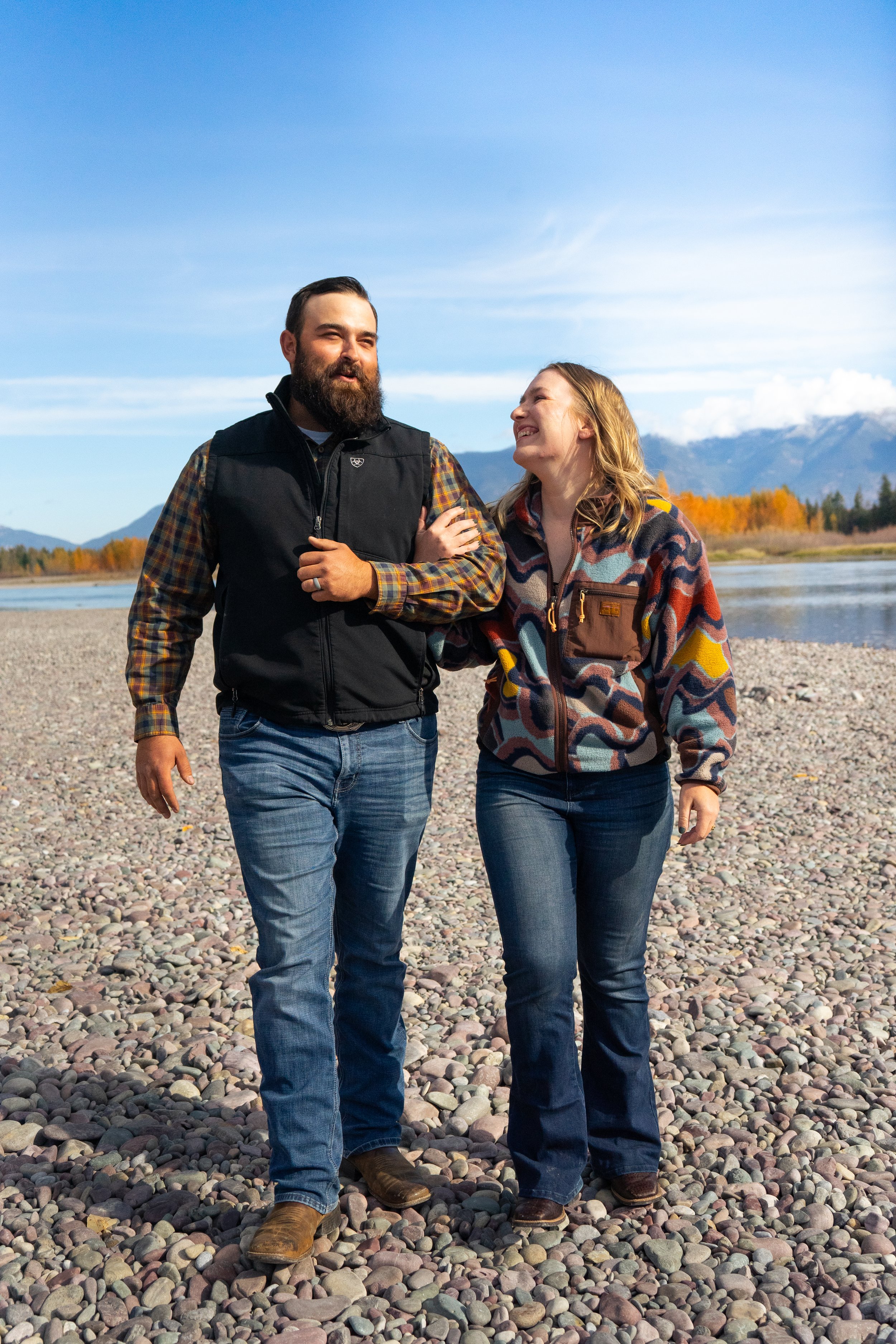 A man and woman walking arm-in-arm on a rocky riverbank with mountains and trees in the background.