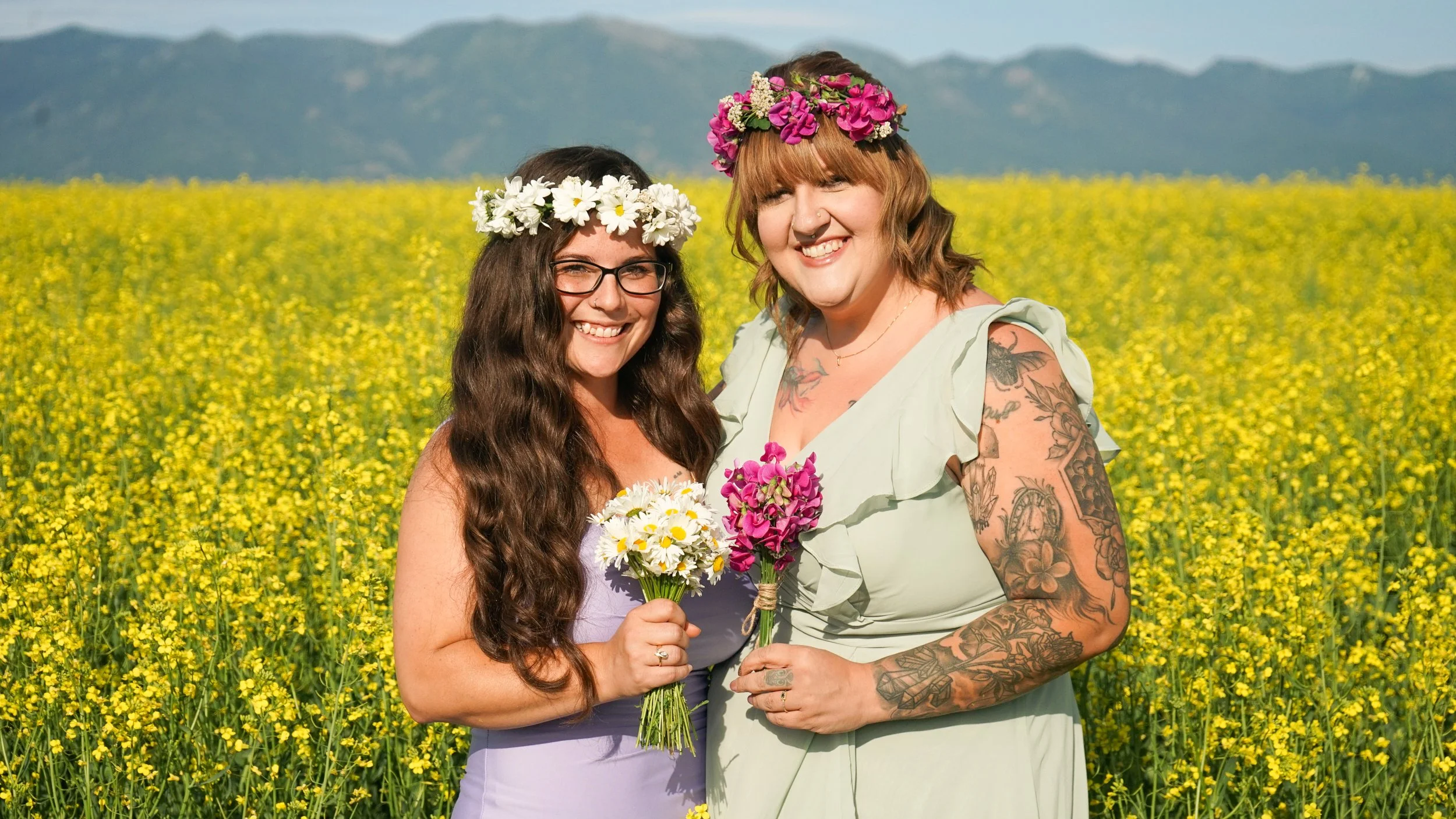 Two women standing together in a yellow flower field, wearing flower crowns and holding bouquets, with mountains in the background, smiling at the camera.