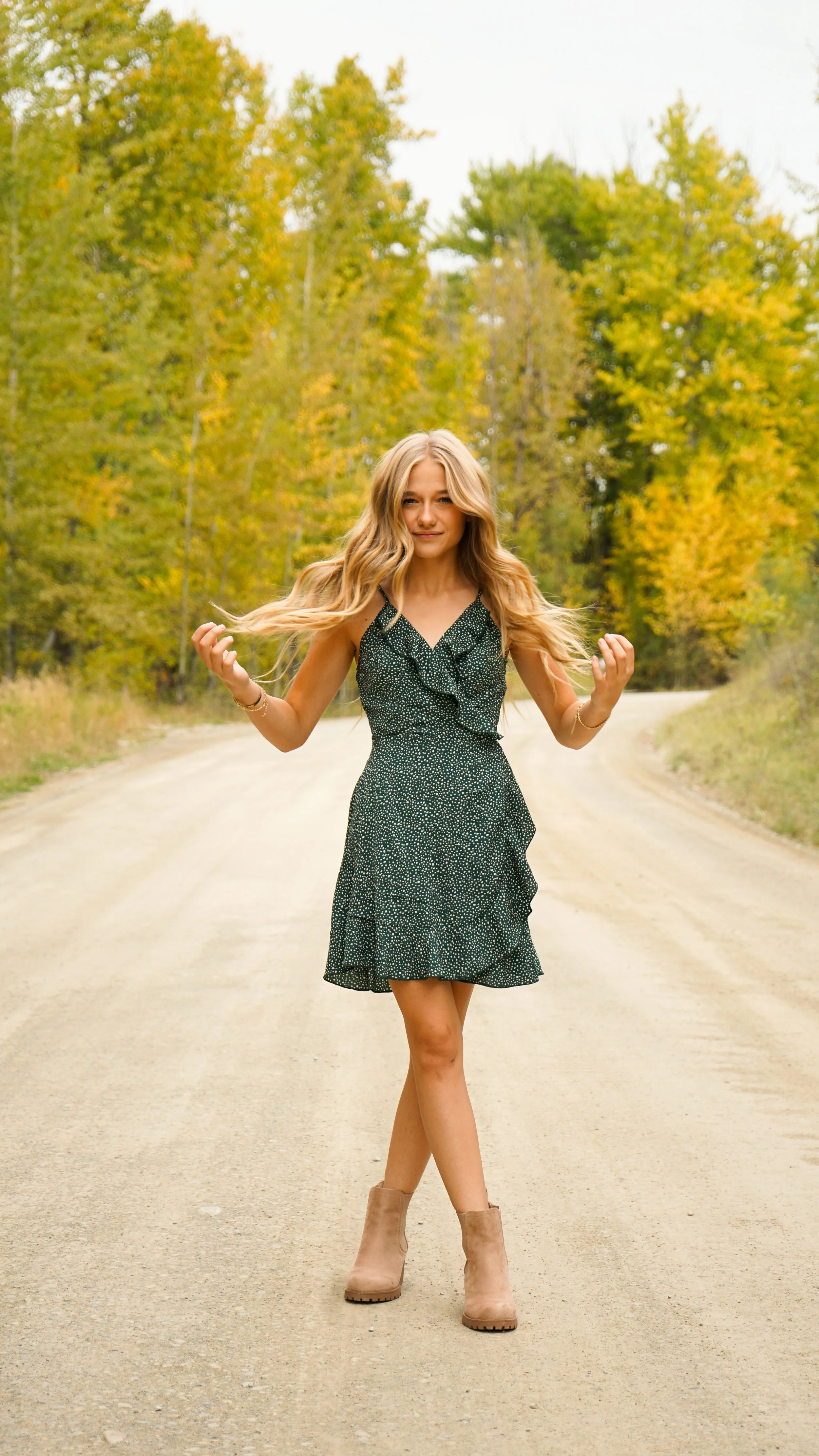 A young woman with long blonde hair in a black polka dot dress and beige boots walking on a dirt road surrounded by forest with green and yellow trees.