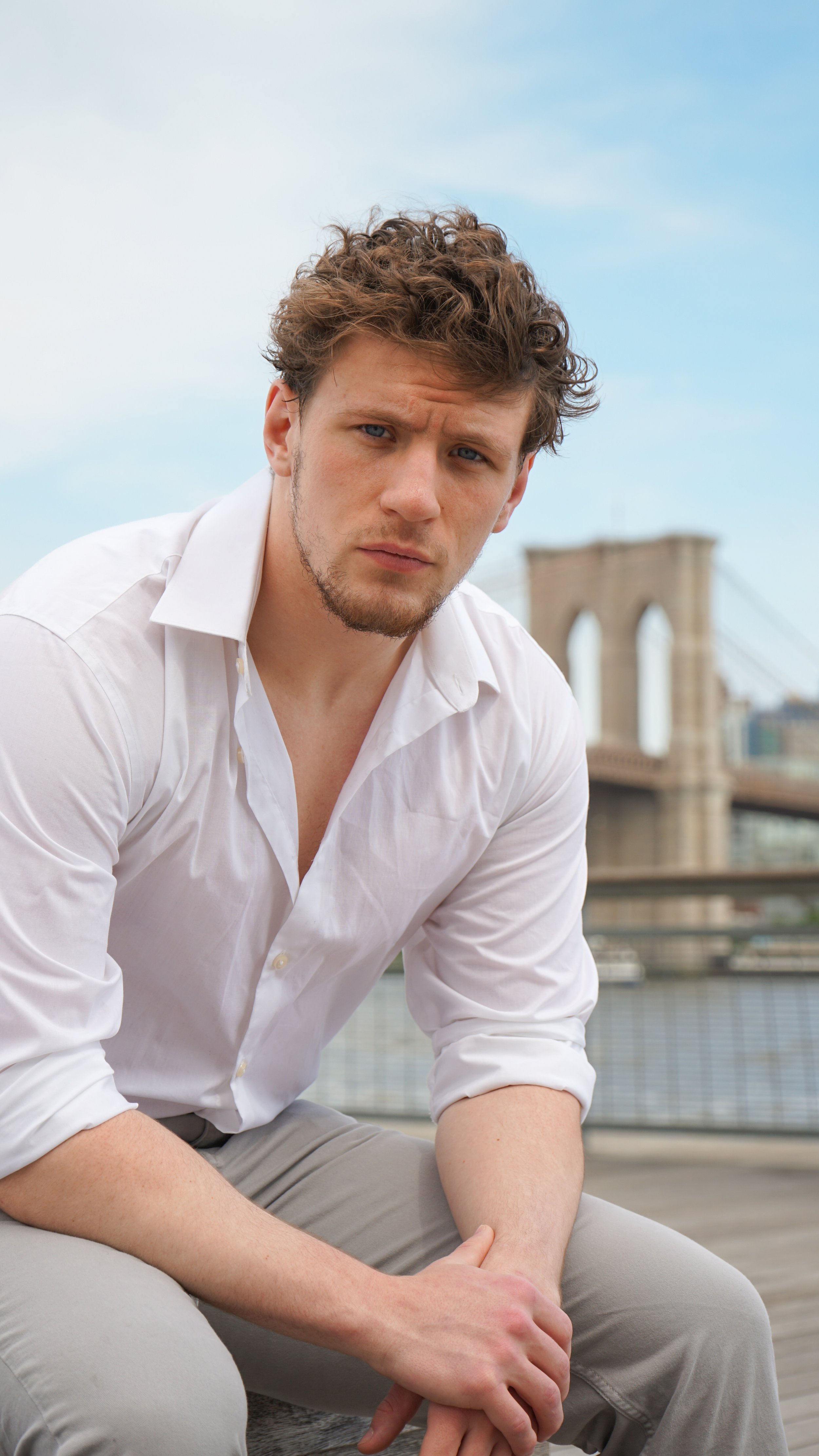 A young man with curly hair and a light beard, wearing a white shirt and beige pants, sitting outdoors with the Brooklyn Bridge in the background.