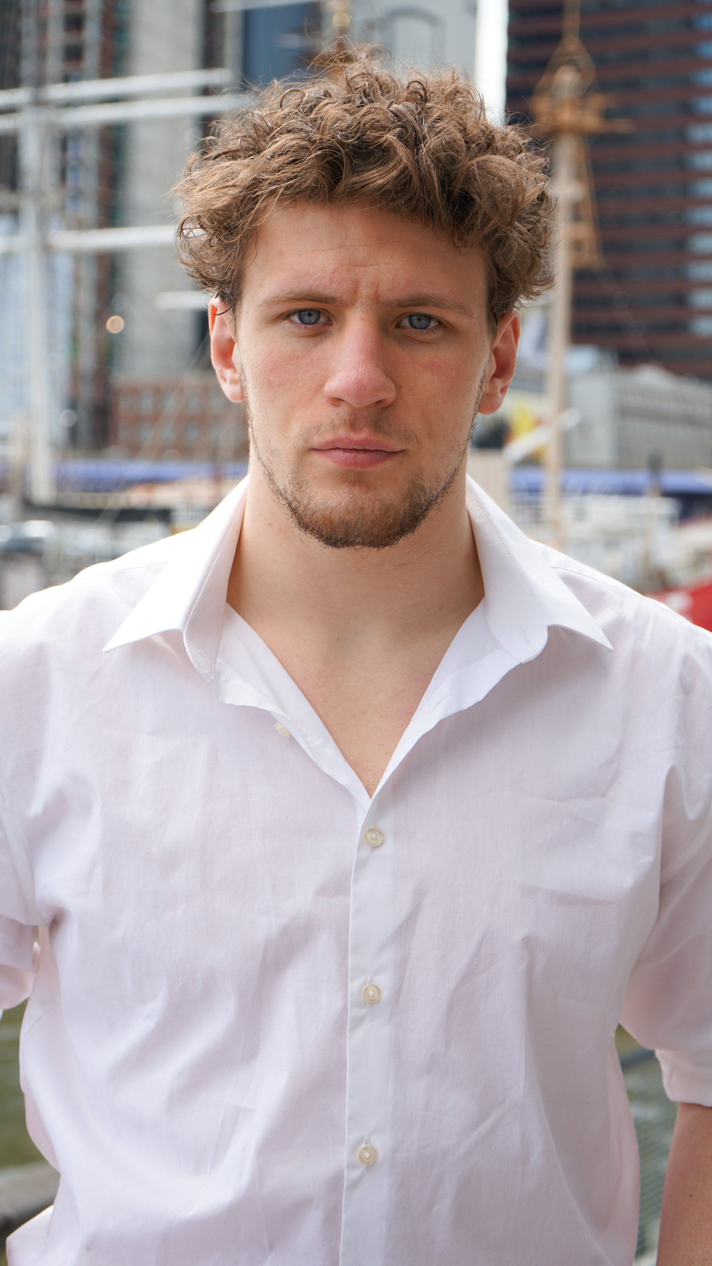 Portrait of a young man with curly brown hair and blue eyes wearing a white shirt, standing outdoors with city buildings and construction in the background.