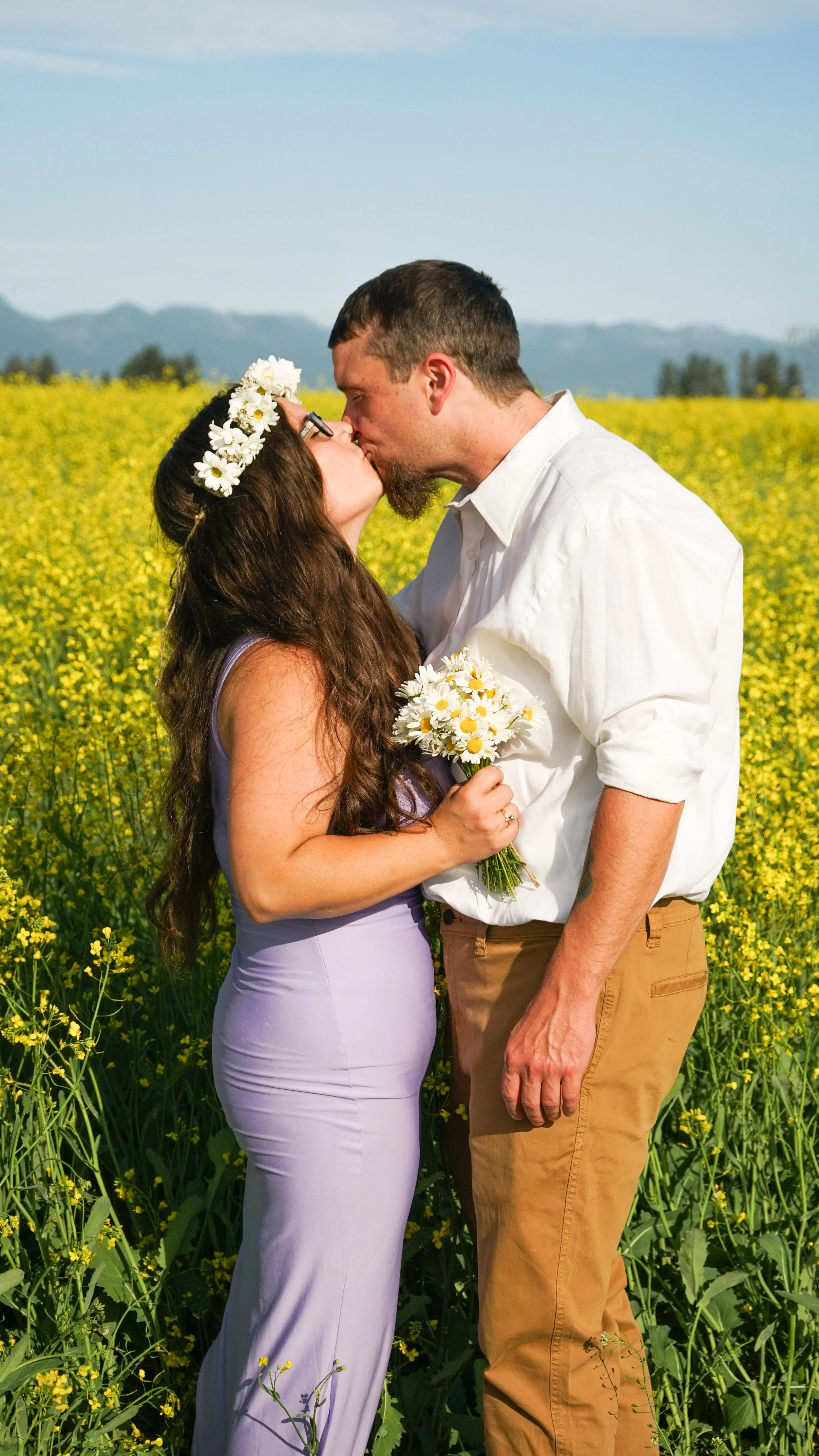 A man and woman kiss in a field of yellow flowers, the woman is holding a bouquet of daisies and wearing a floral crown; man wears a white shirt and tan pants.