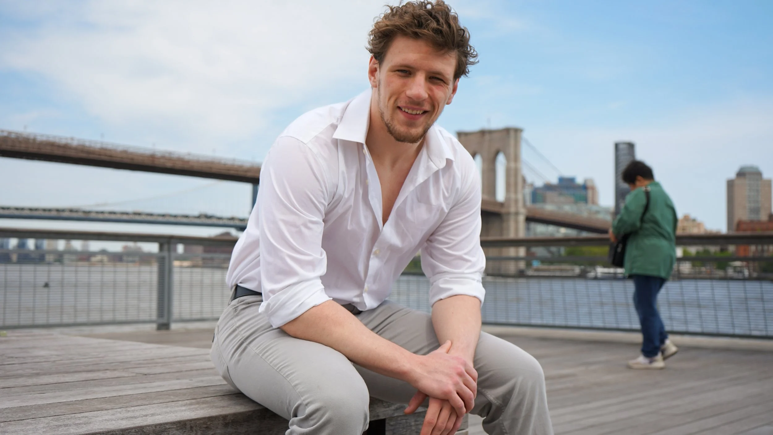 A young man with curly brown hair and light skin, wearing a white shirt and light-colored pants, sitting on a wooden bench outdoors near the Brooklyn Bridge in New York City. A woman with dark hair, wearing a green jacket, is walking in the backgroun