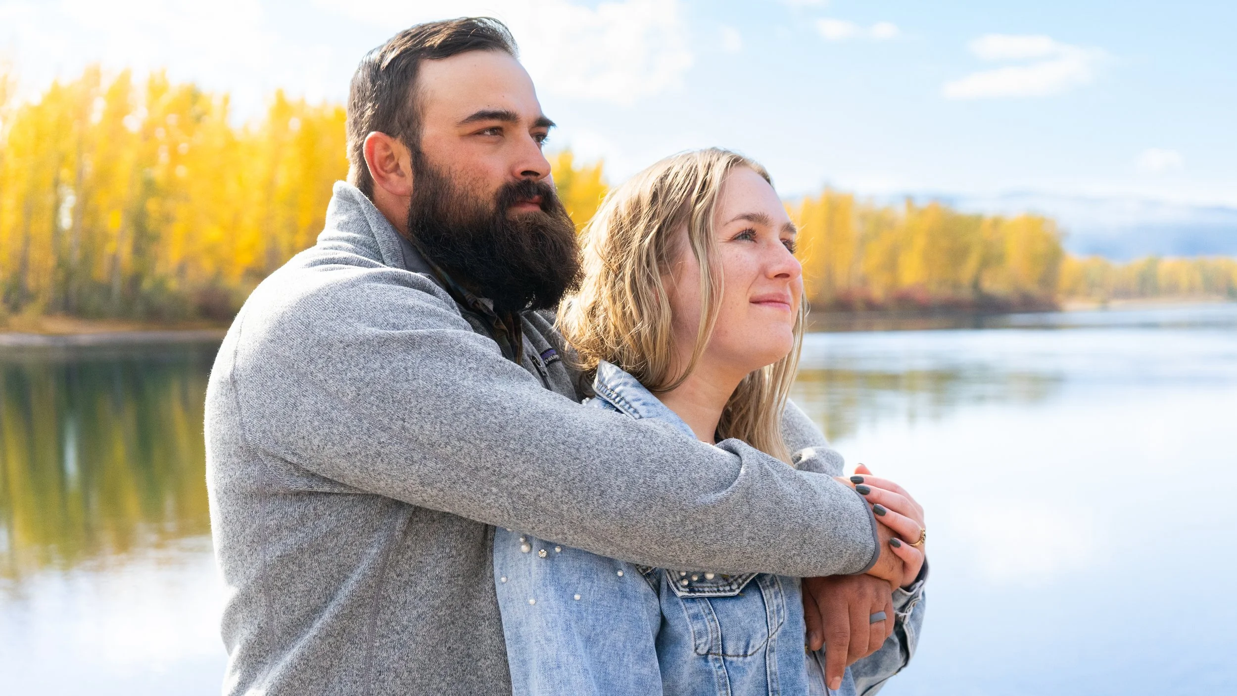 A man with a beard and a woman with blonde hair standing by a body of water, with trees displaying fall foliage in the background.