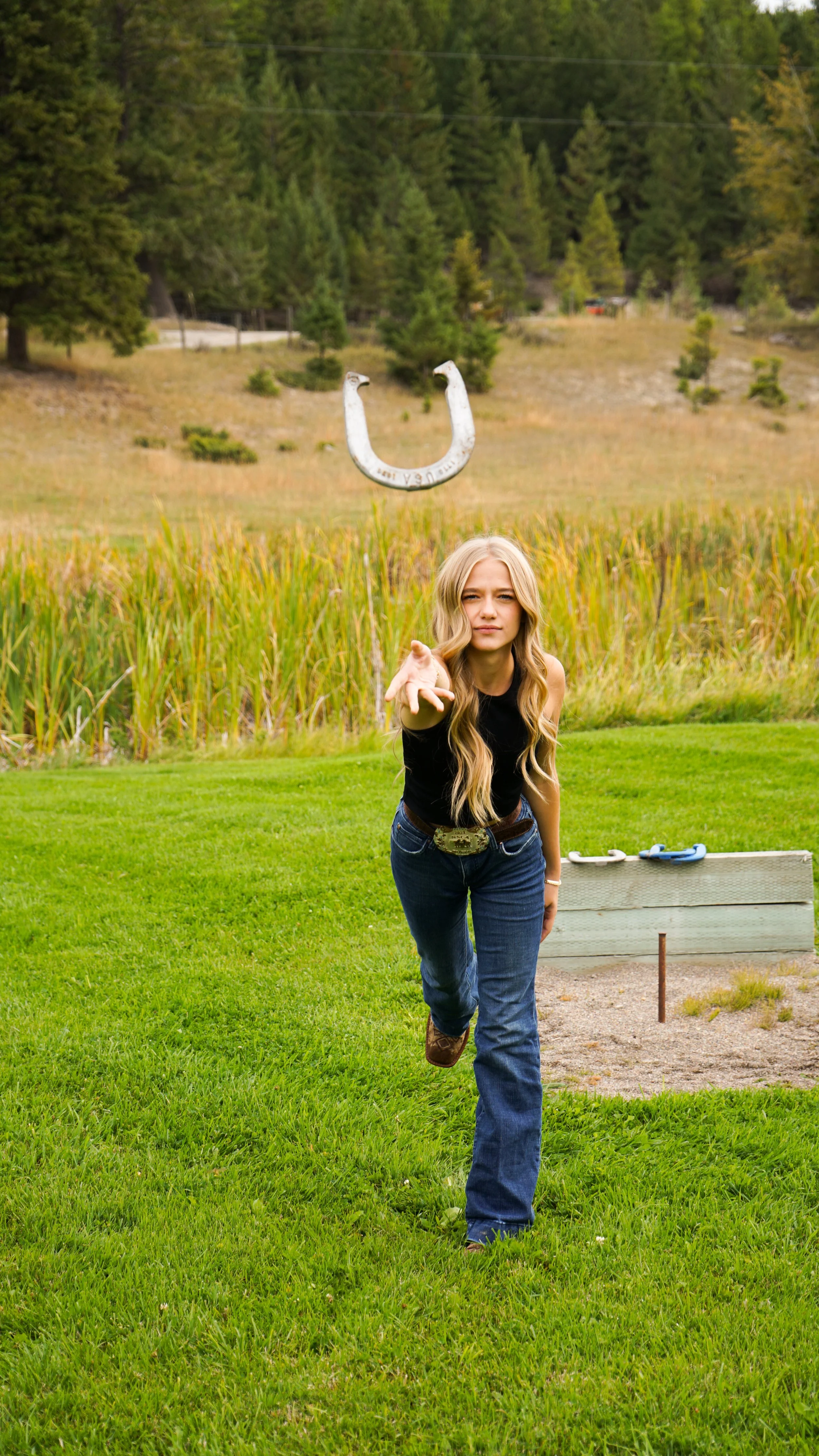 Young woman in jeans and a black top throwing a horseshoe outdoors in a grassy field, with trees and a horseshoe game station in the background.