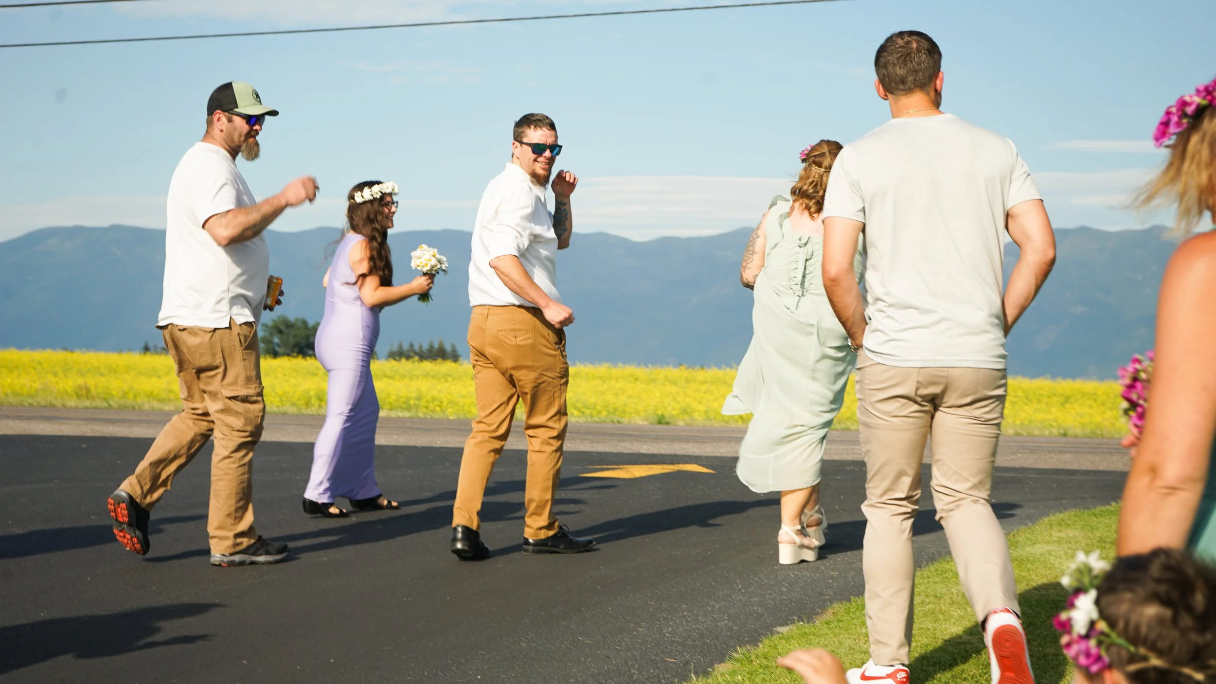 A group of people, including men and women, standing and walking on a rural road during daytime, with mountains and a clear sky in the background.