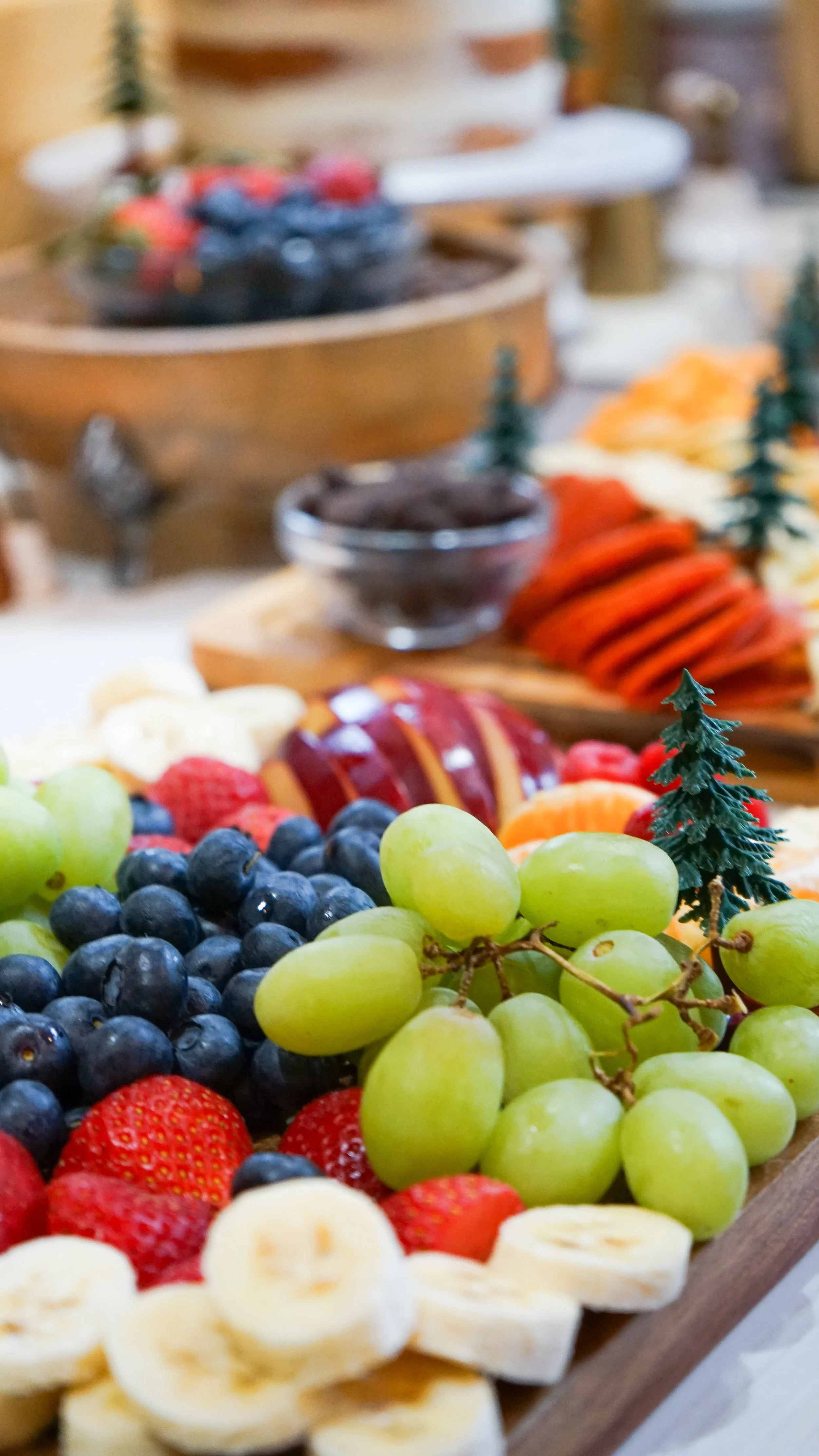 A variety of fresh fruits on a wooden platter, including green and black grapes, strawberries, sliced bananas, and other assorted fruits in the background.