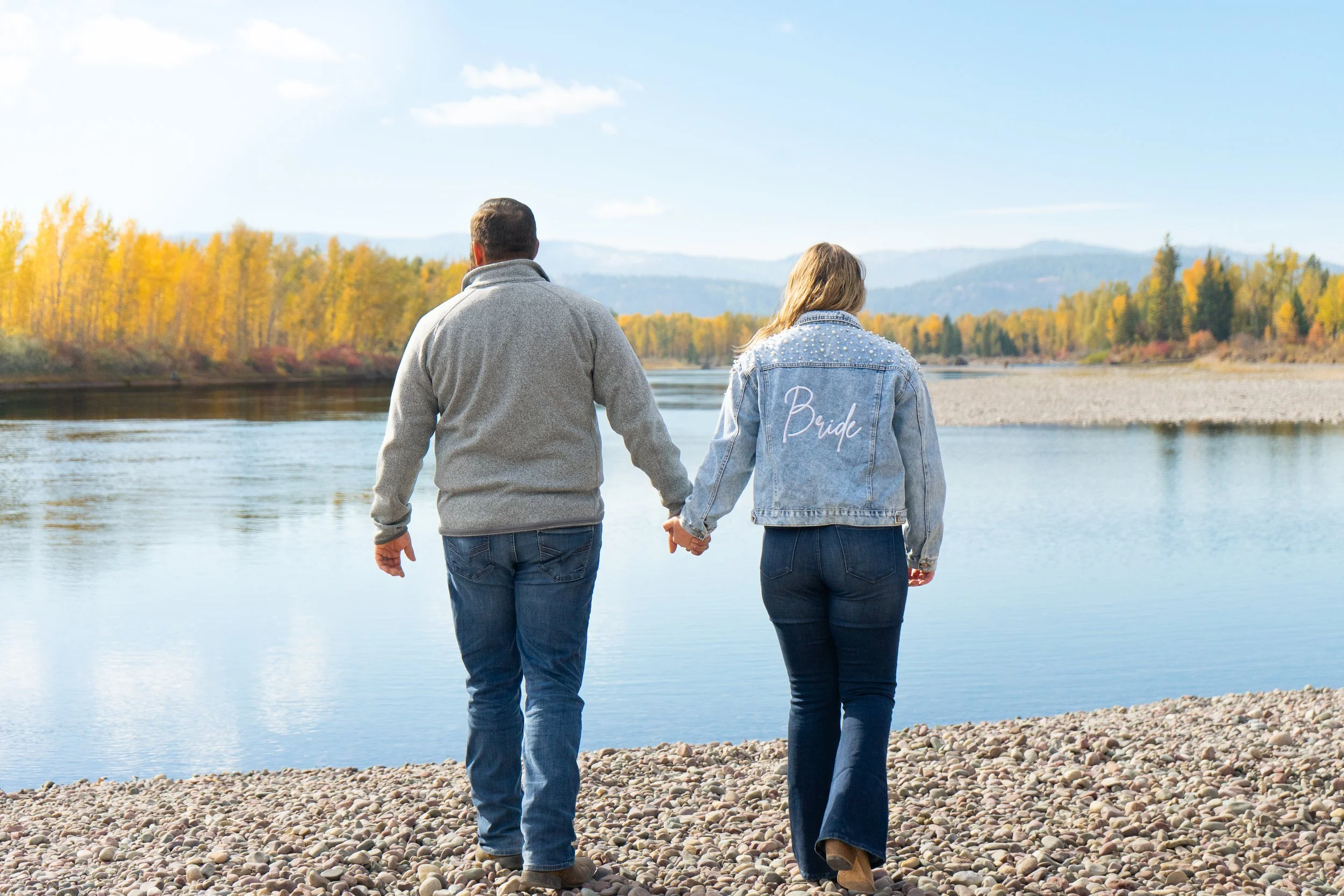 A couple holding hands walks by a lake with autumn trees in the background, one woman wearing a denim jacket with "Bride" written on the back.