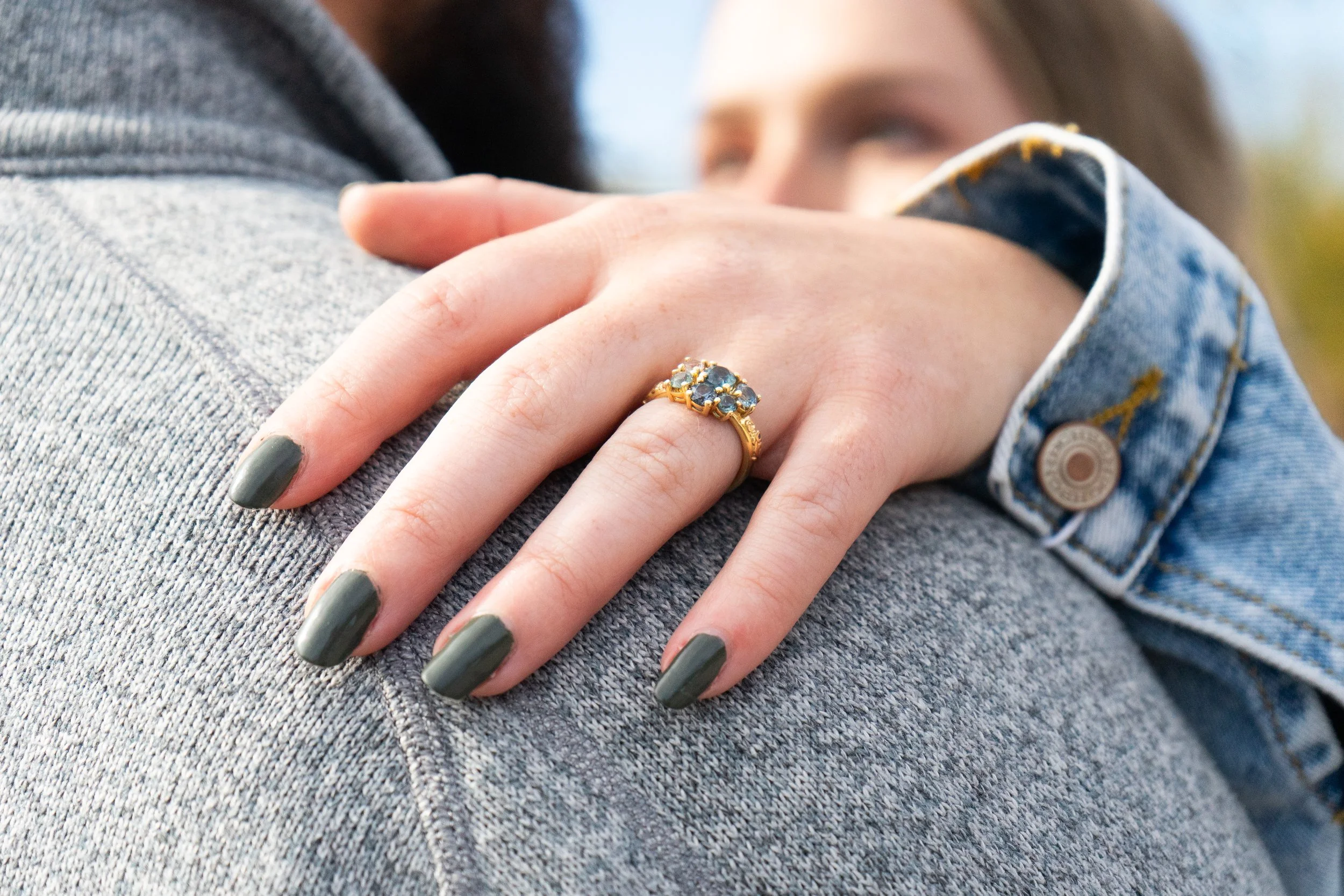 A close-up of a person wearing a gold ring with blue stones hugging another person, who is wearing a gray sweater and a denim jacket.