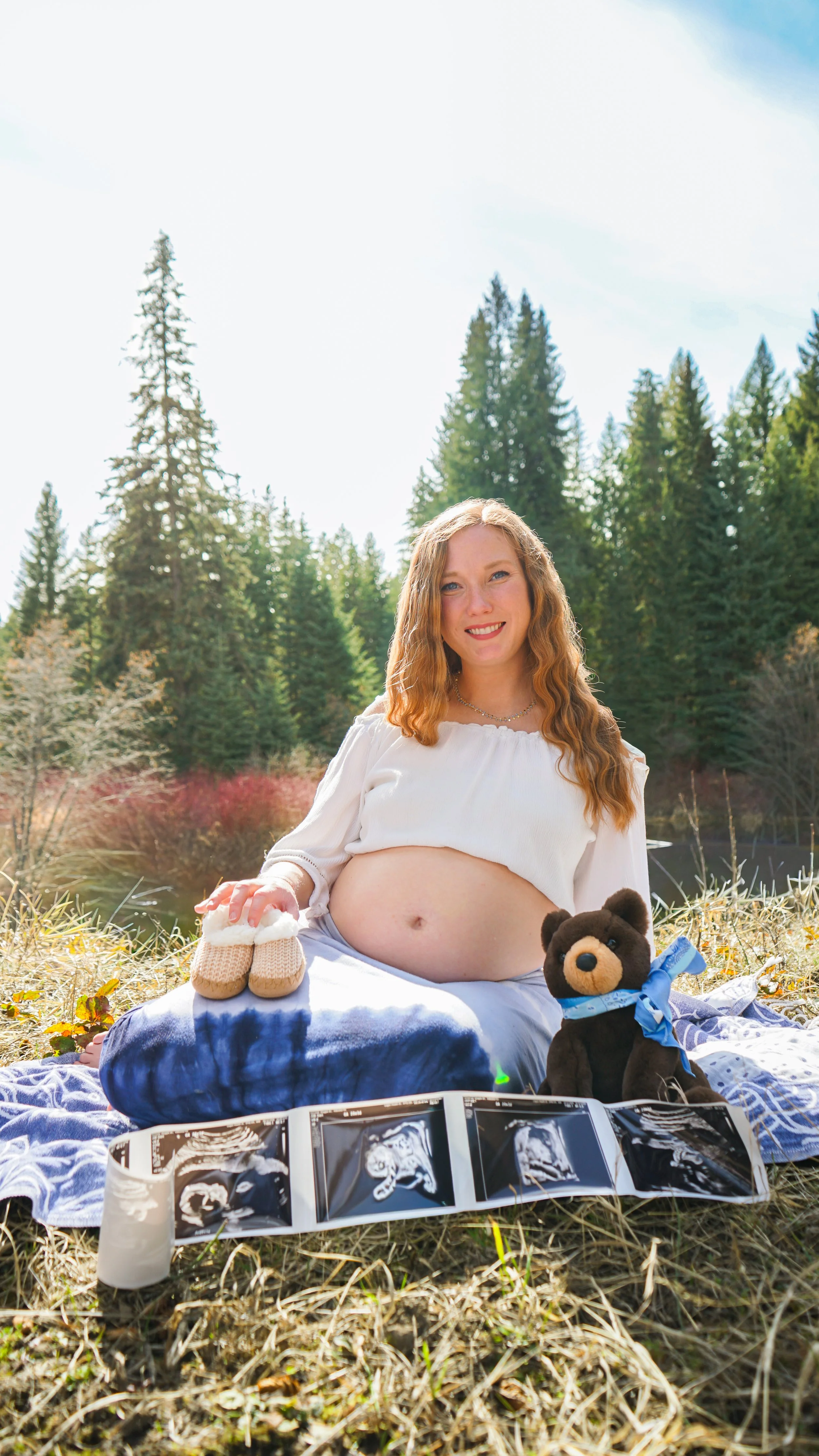 A pregnant woman sitting outdoors on a blanket in a forested area during daytime, with ultrasound images displayed in front of her, and stuffed animals including a teddy bear and baby shoes nearby.