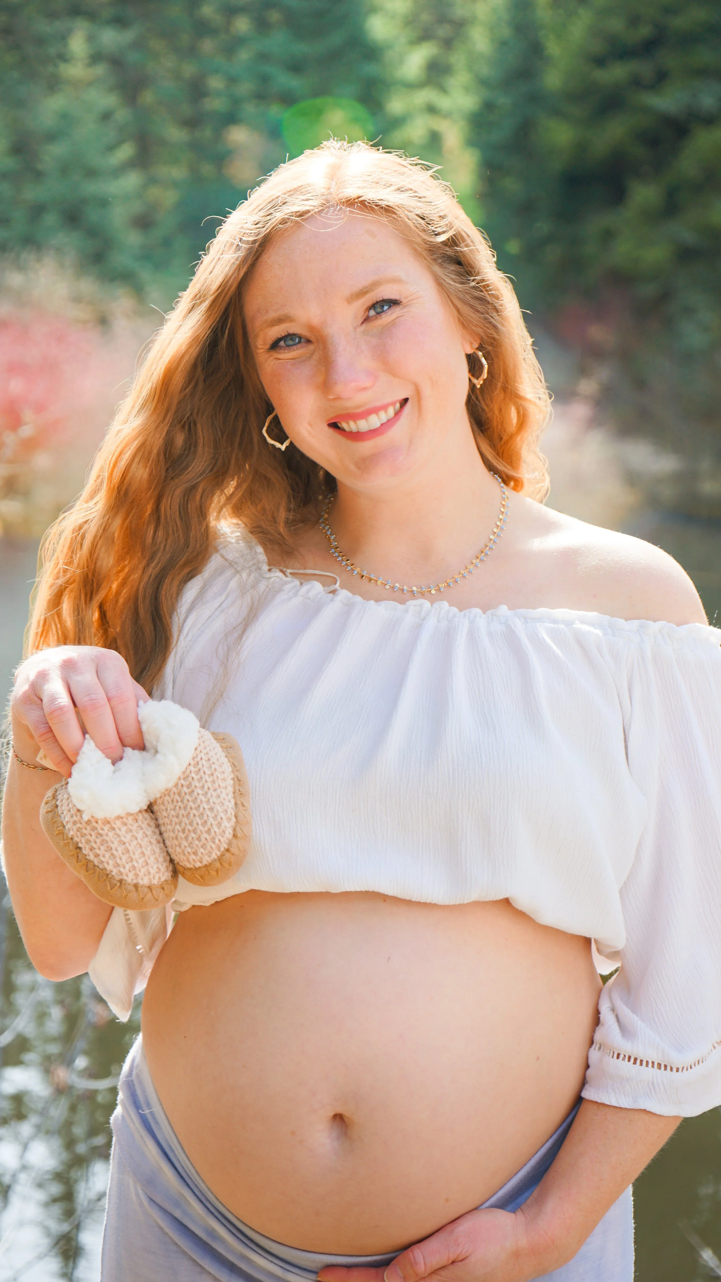 A pregnant woman with long red hair, smiling, holding her belly, outdoors, with water and trees in the background.