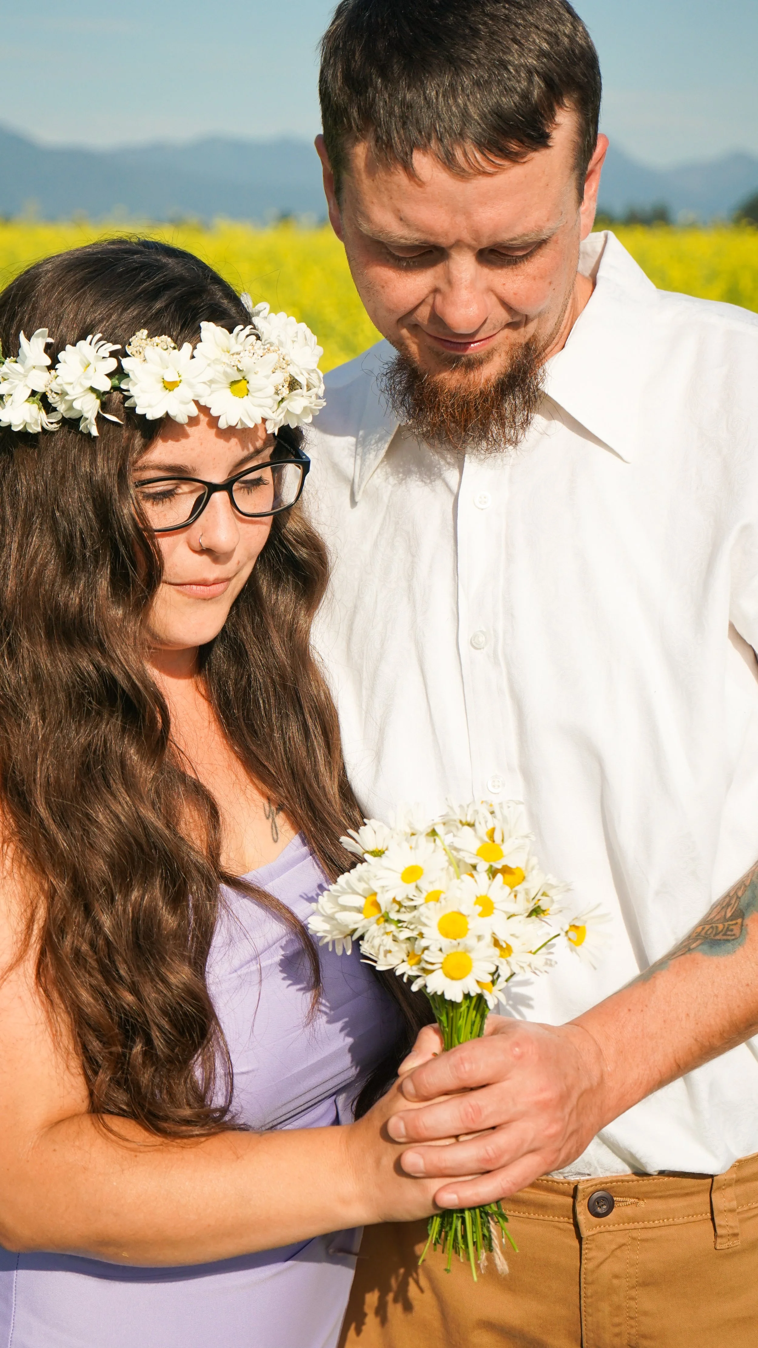 A woman with long brown hair, glasses, a flower crown, and a purple dress holds a bouquet of daisies. A man with a beard, white shirt, and tan pants stands beside her, holding the bouquet along with her. They are outdoors in a field with yellow flowe