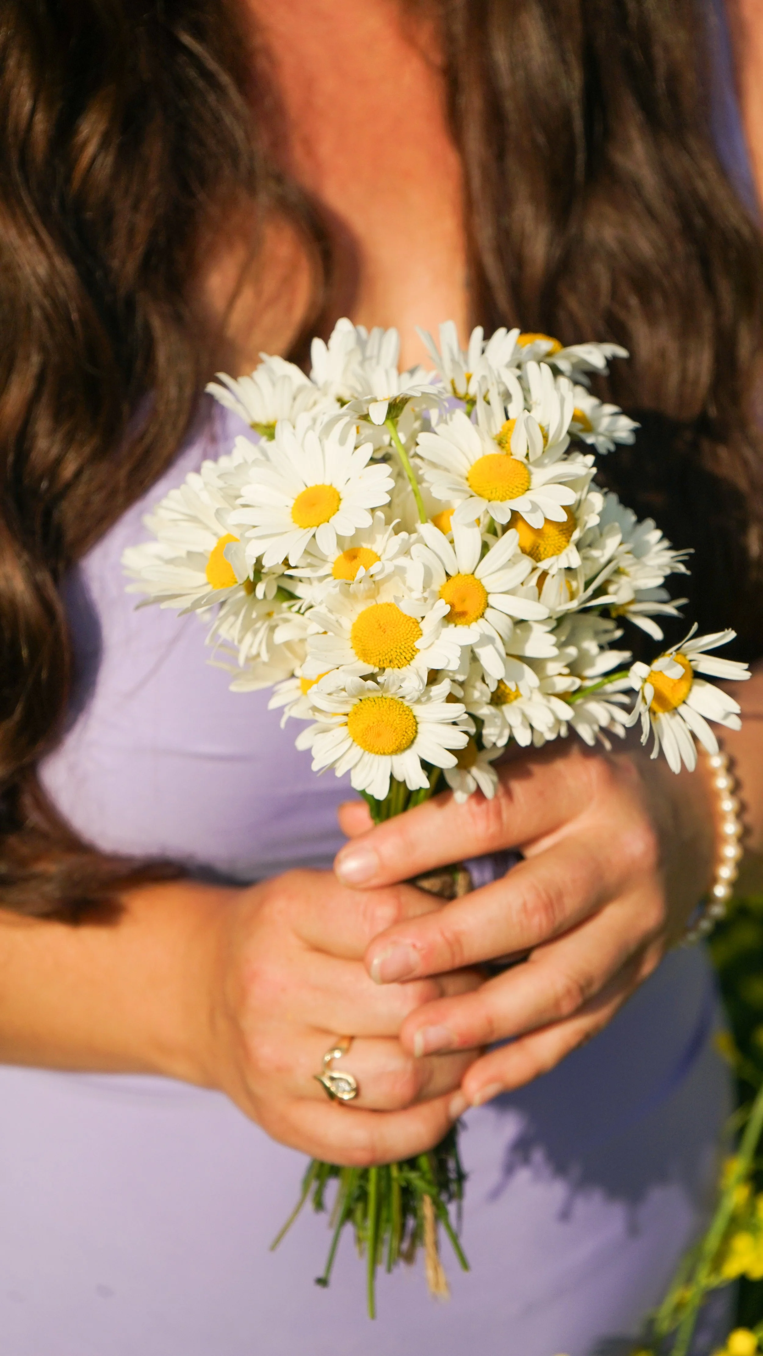 A woman with long, dark, wavy hair holding a bouquet of daisies with white petals and yellow centers.