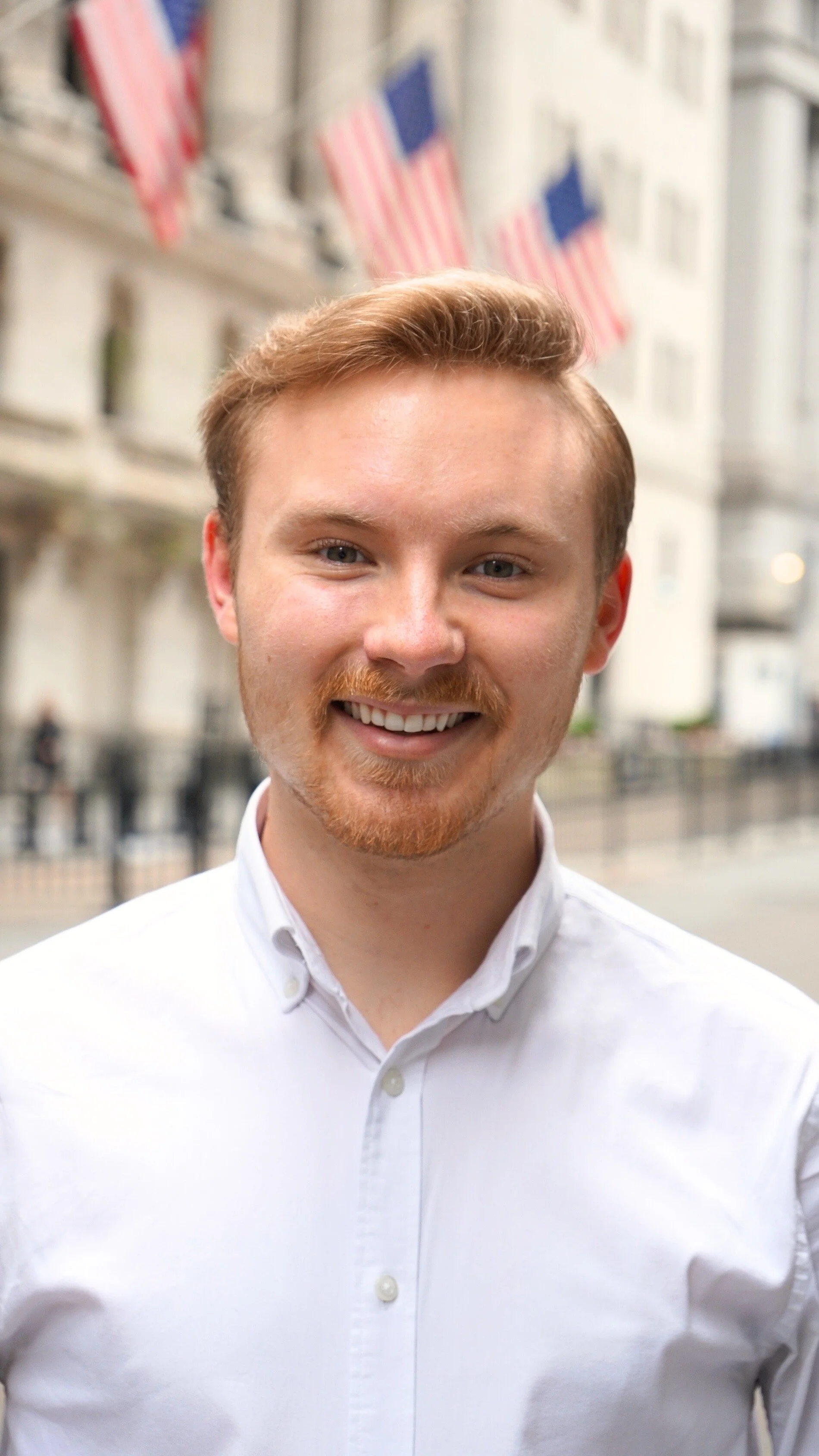 A young man with light brown hair and a beard, smiling while standing outside in front of a building with American flags.