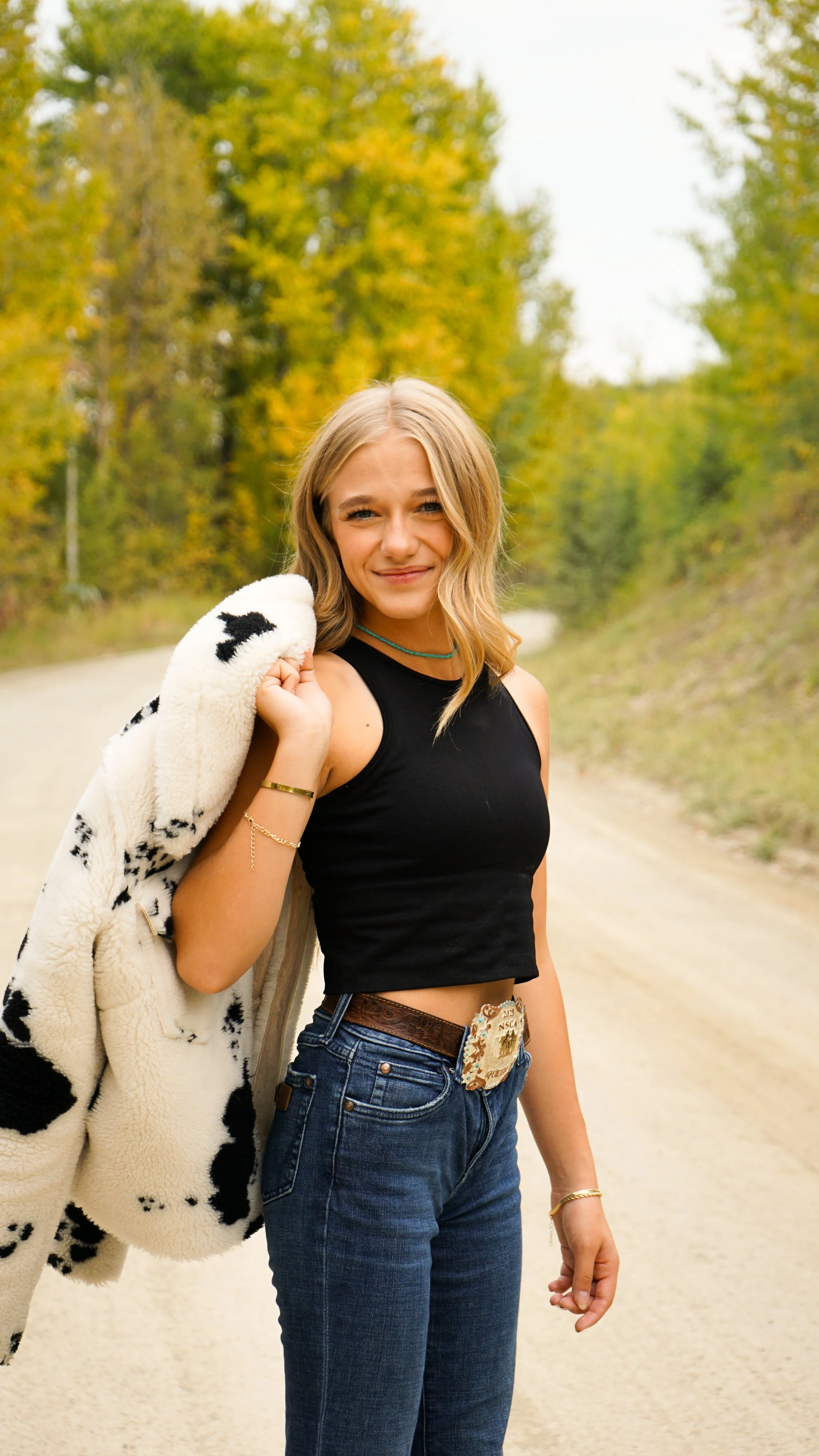 A young woman standing on a dirt road surrounded by trees with fall foliage, wearing a black crop top and jeans, carrying a white and black patterned fleece jacket over her shoulder, smiling at the camera.