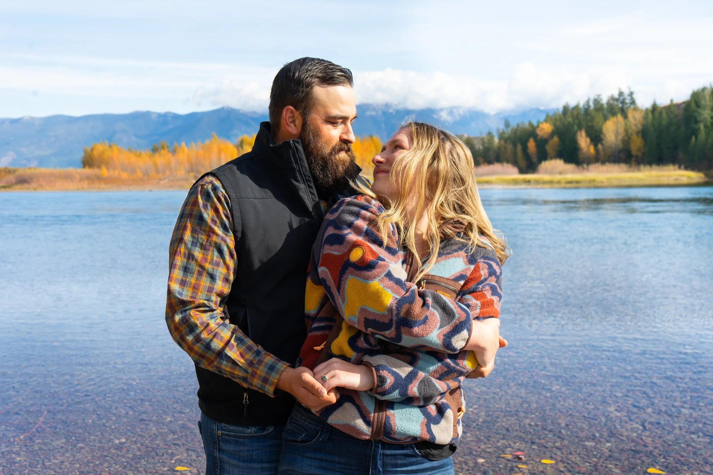 A couple standing by a lake, embracing each other with mountains and trees in the background during fall.