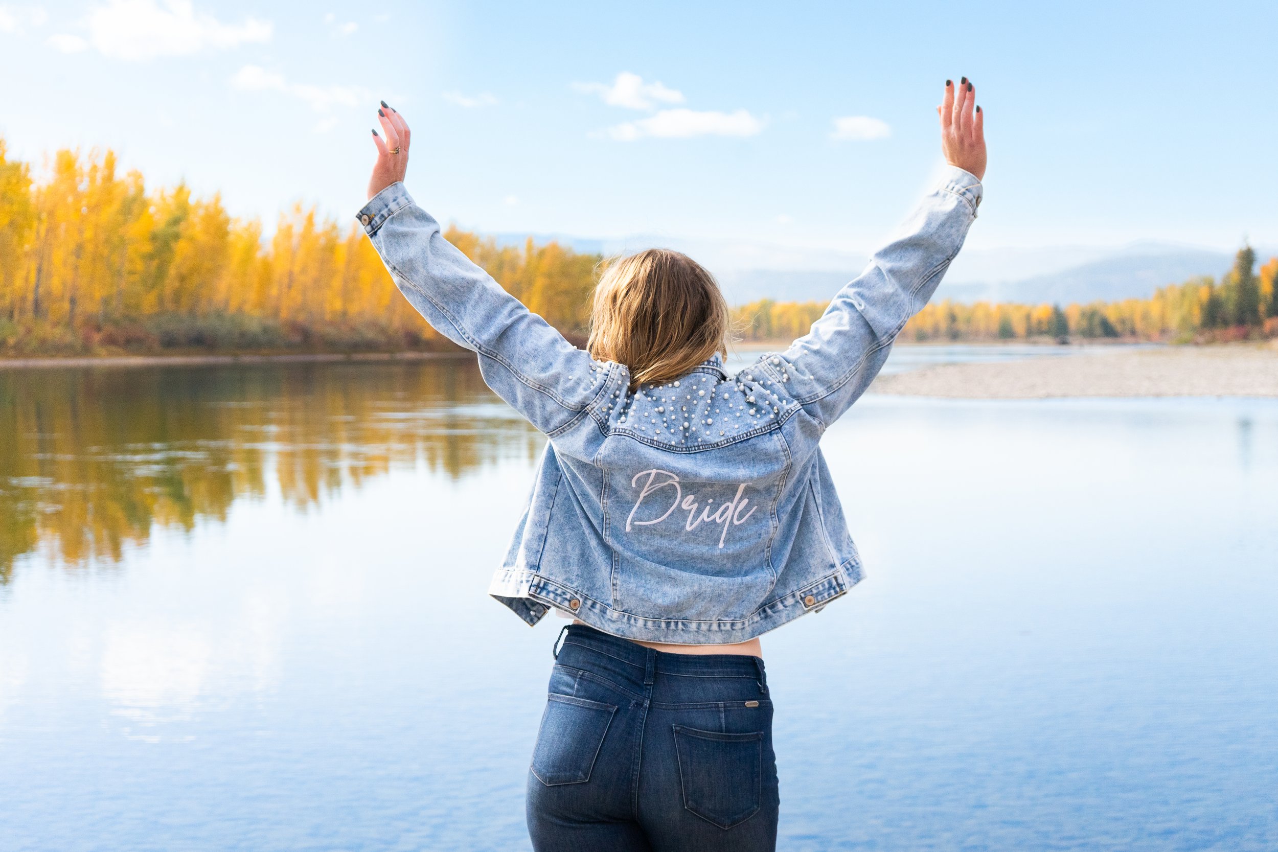 A person with long hair wearing a denim jacket with 'Bride' embroidered on the back, standing by a lake with arms raised, trees with autumn leaves in the background, and a clear blue sky.