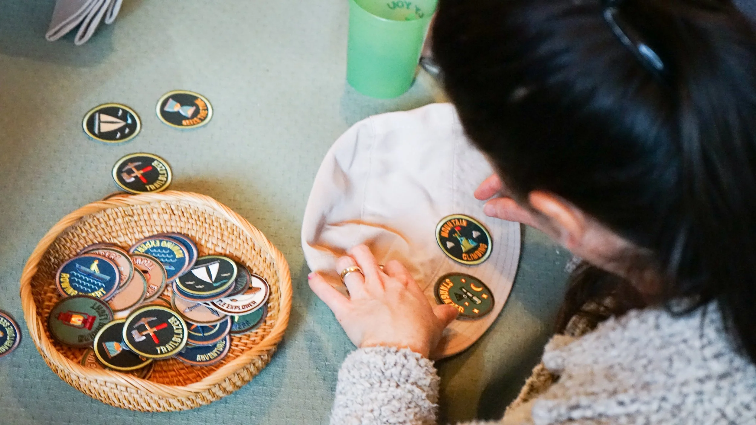 A person with dark hair and a fuzzy beige sweater places a round patch onto a white shirt with a Scooby-Doo patch on it. There are several patches with various designs and words like "Travel," "Adventures," and "Joy" and a basket filled with more pat