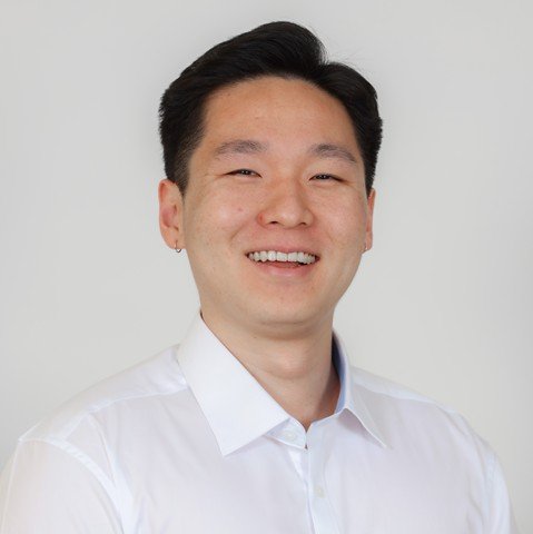 A smiling man with short black hair wearing a white dress shirt against a plain background.