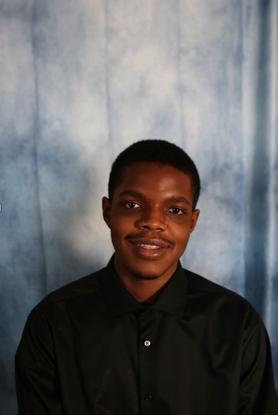 A young man is smiling and wearing a black shirt, standing in front of a blue curtain.