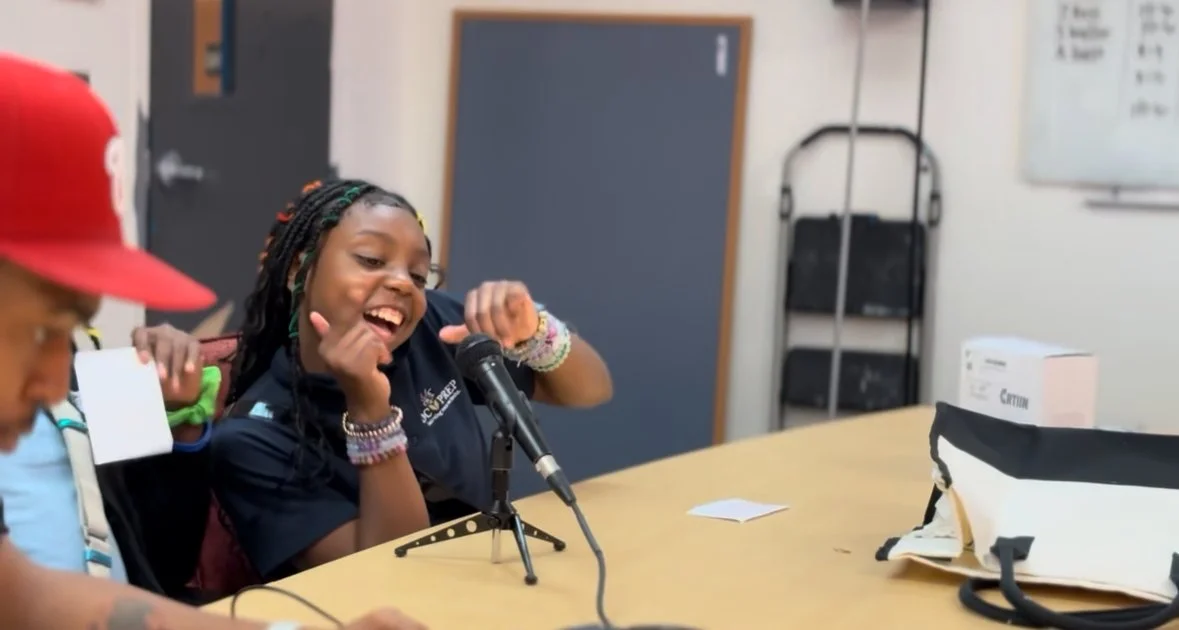 A young girl with braided hair and colorful bracelets speaking into a microphone during a radio or podcast interview, seated at a table with other children nearby, in a room with a chalkboard and office supplies.