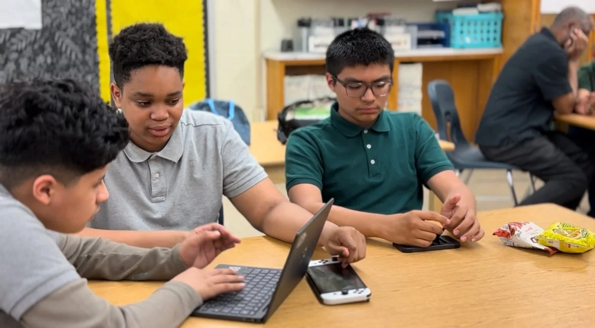 Three boys sitting at a table in a classroom, using electronic devices, with another person in the background.