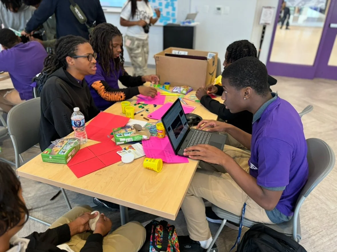 Group of students sitting at a table working on a craft project with colorful paper, playdough, and a laptop, in a school setting.