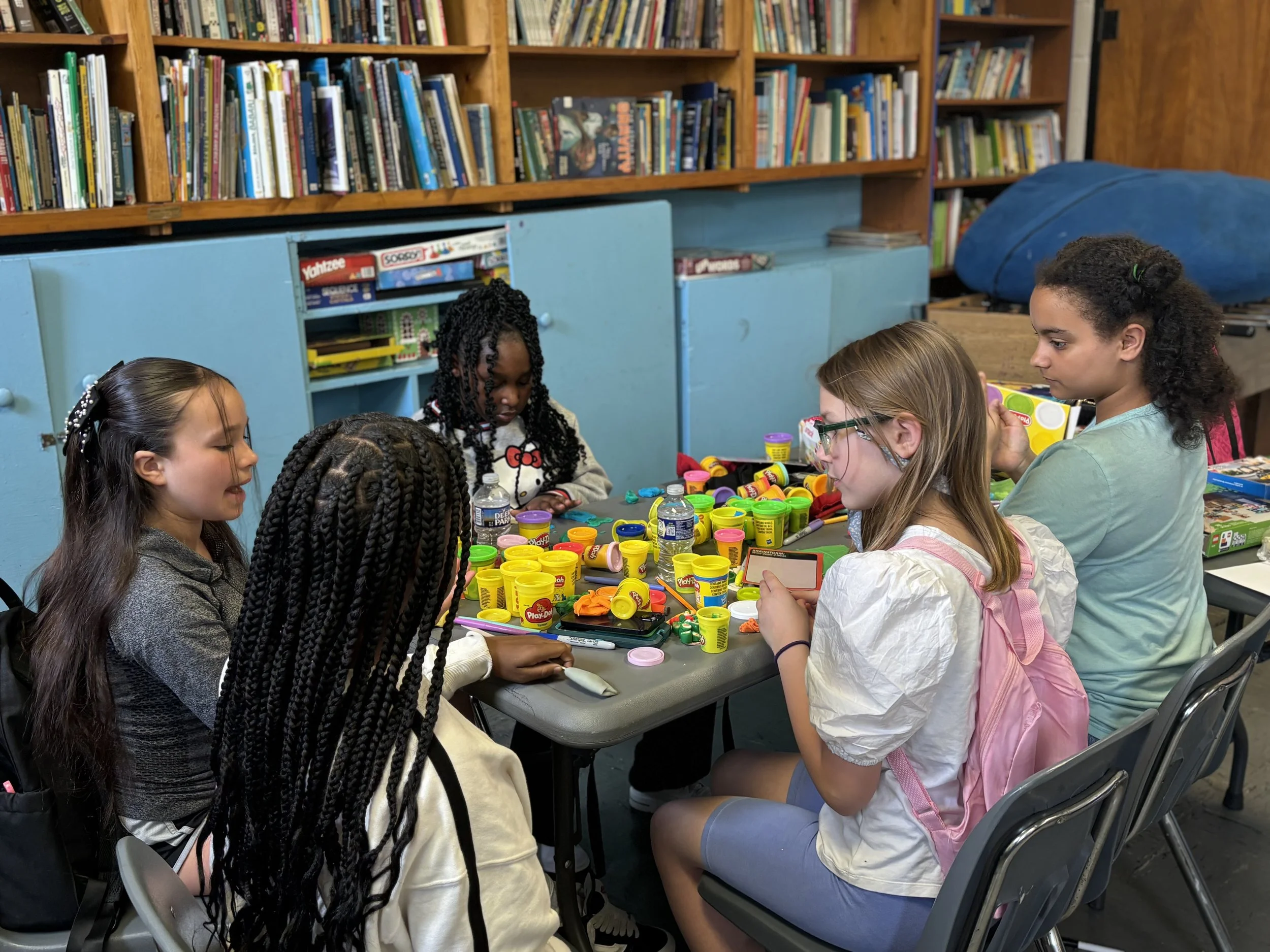 Six children sitting at a table playing with colorful Play-Doh in a room with bookshelves and toys.