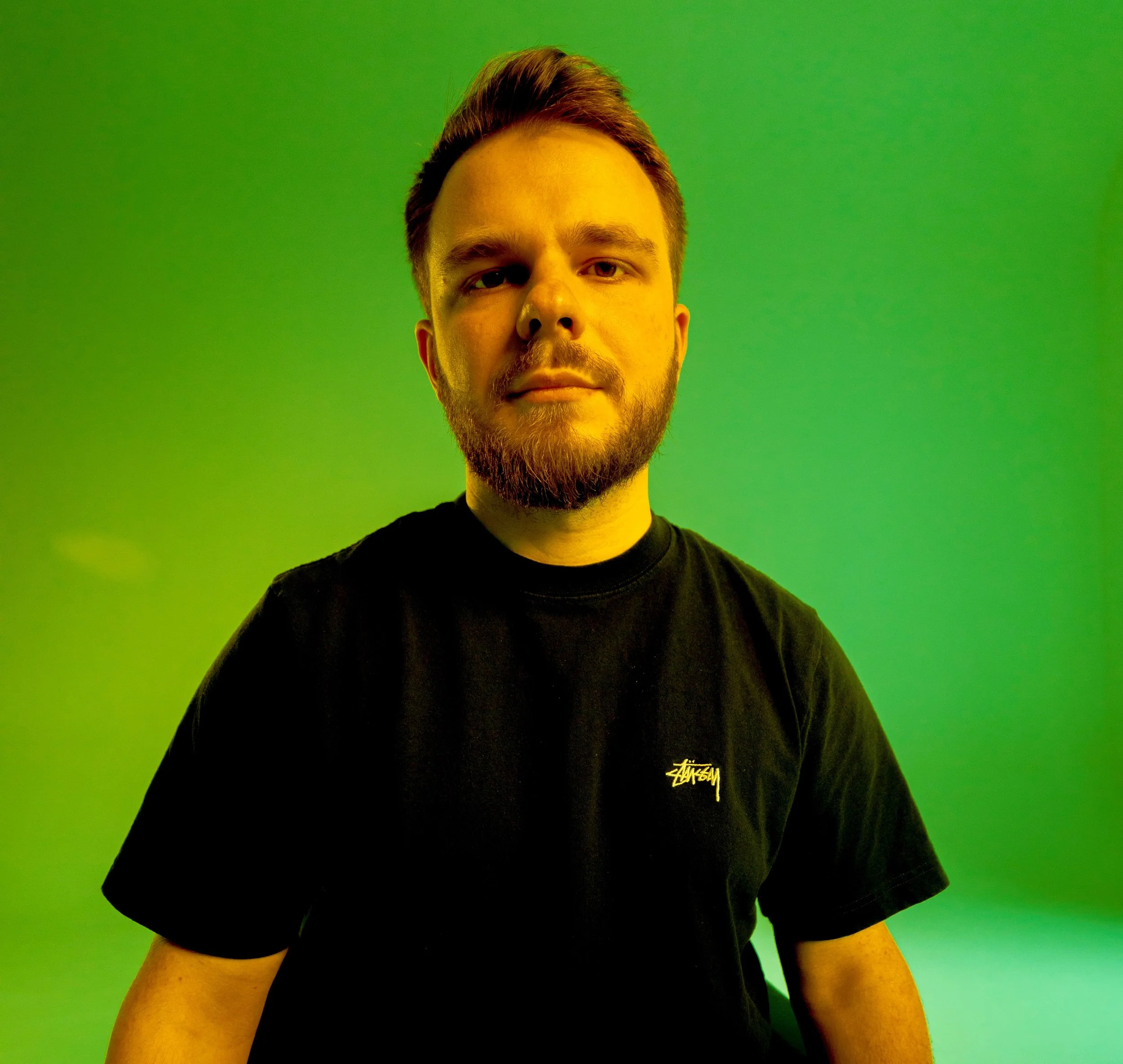A young man with a beard and styled hair, wearing a black T-shirt, stands against a green background.