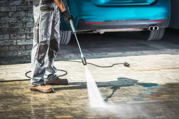 A person wearing gray work pants and brown boots using a pressure washer to clean a brick driveway next to a blue car.