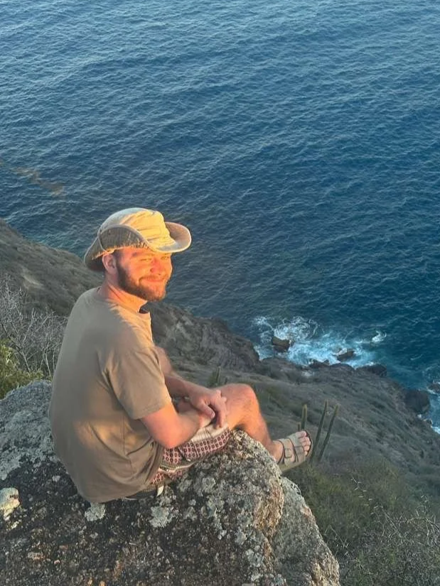 A man sitting on a rocky cliff overlooking the ocean, smiling, wearing a beige shirt, shorts, sandals, and a bucket hat.