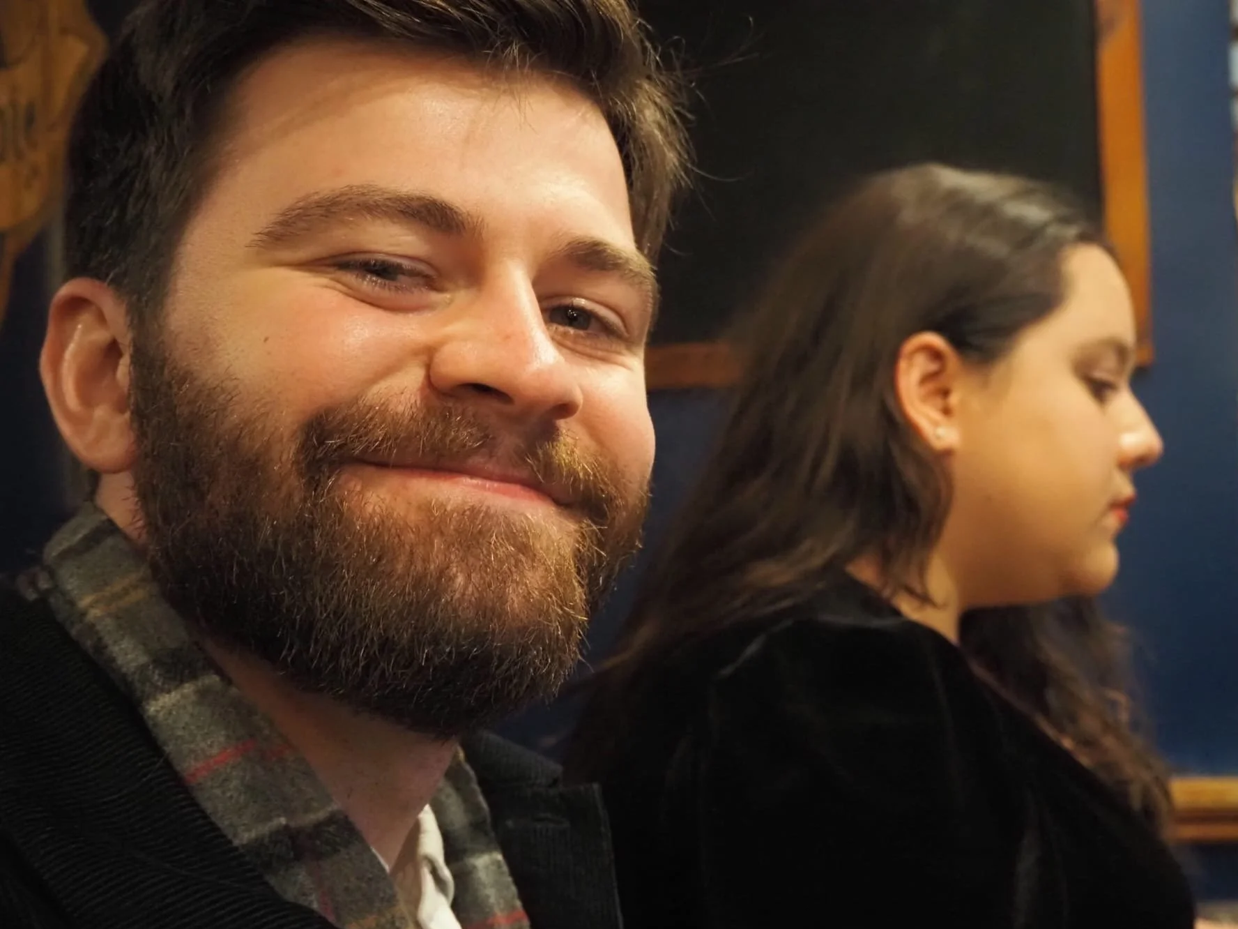 Close-up photo of a smiling man with a beard and short brown hair sitting next to a woman with long dark hair, both facing sideways, in a dimly lit room with a black and blue wall.