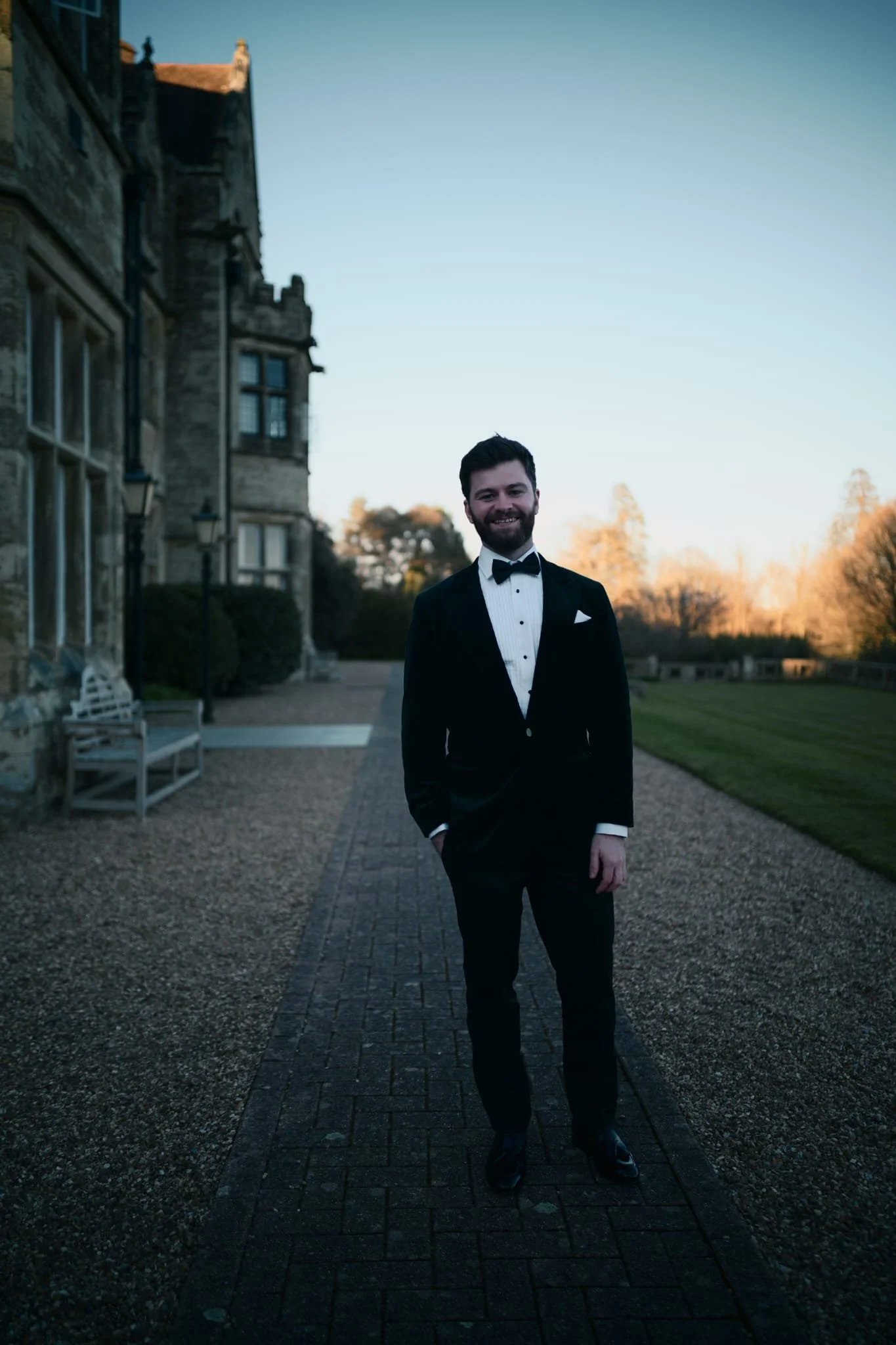 A man in a tuxedo standing outside on a cobblestone path in front of an old stone building at dusk